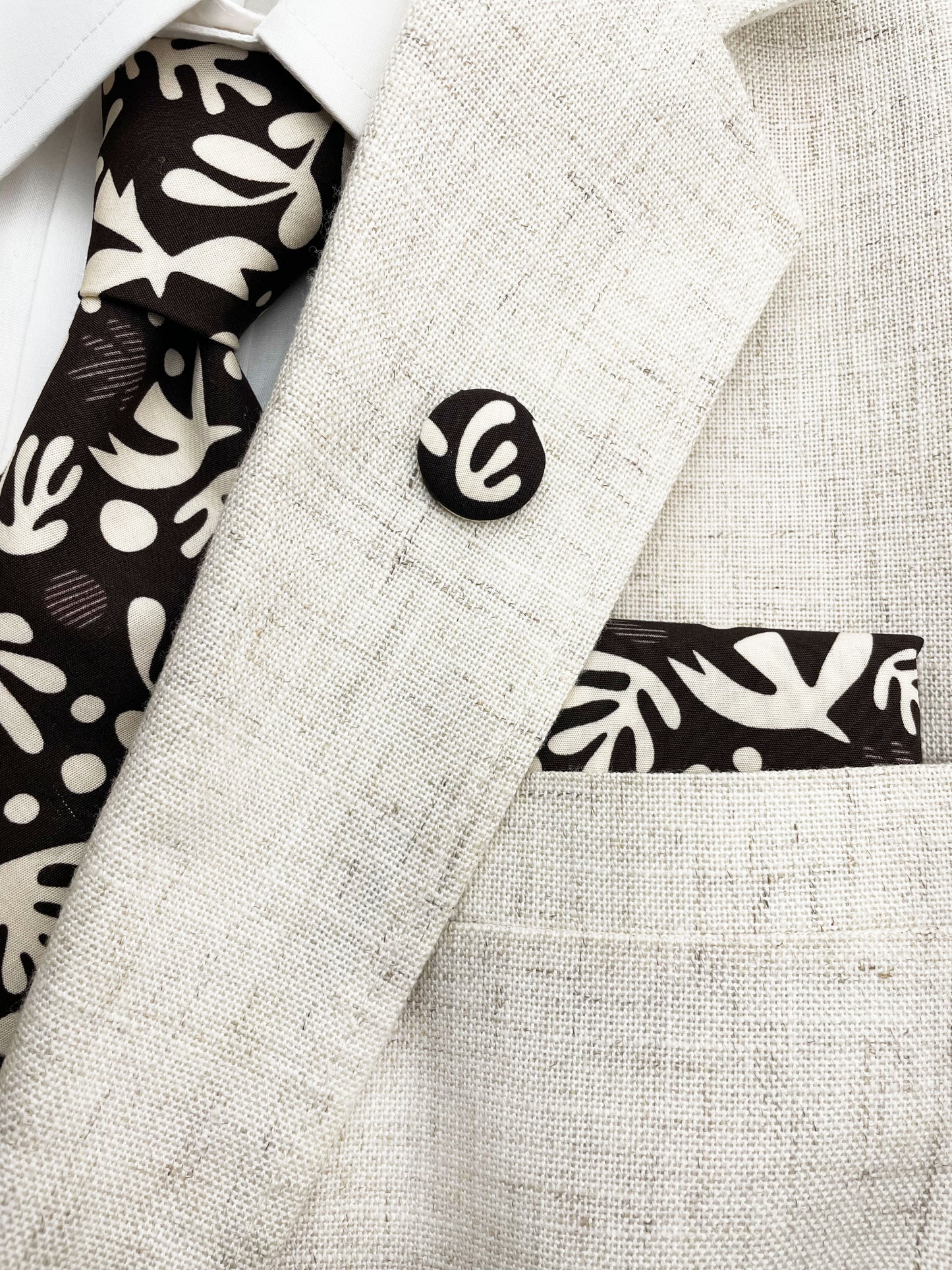 A cotton necktie with cream-colored birds and abstract shapes shown with a matching pocket square and lapel pin.