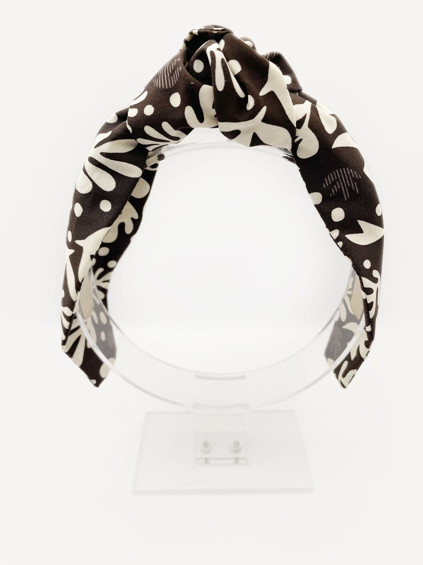 A dark charcoal gray knotted headband with cream-colored birds and abstract shapes. The headband is displayed on an acrylic stand.