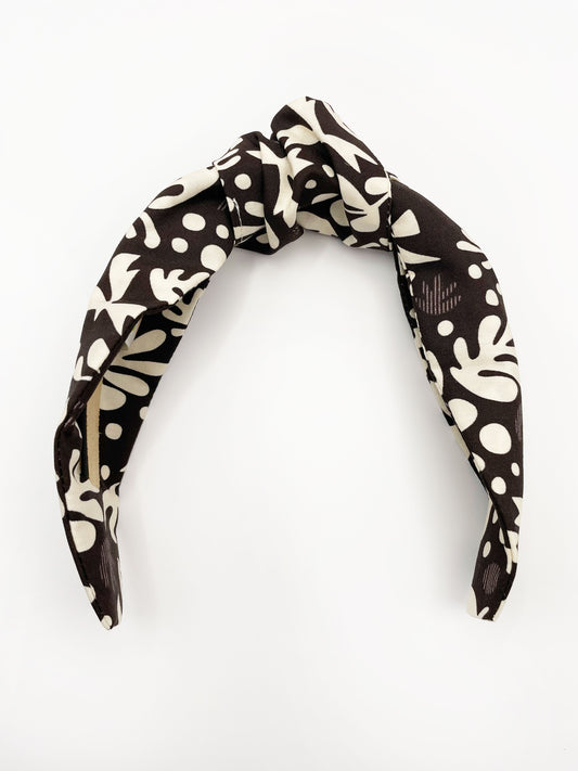 A dark charcoal gray knotted headband with cream-colored birds and abstract shapes. Each headband is crafted with cotton fabric attached to a durable hair band for a secure and comfortable fit.