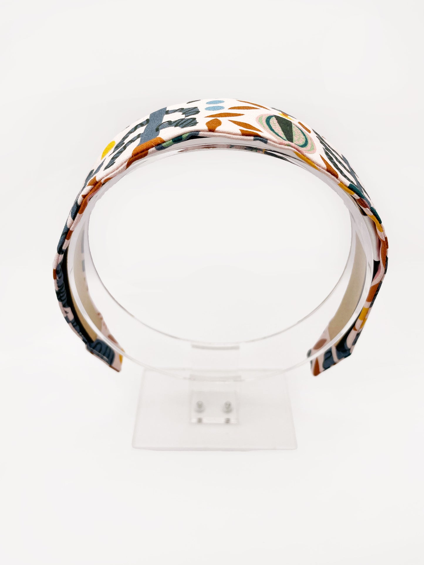A fabric headband featuring a bold nature-inspired print of bugs, plants, suns, and rainbows. The headband is shown on an acrylic display.