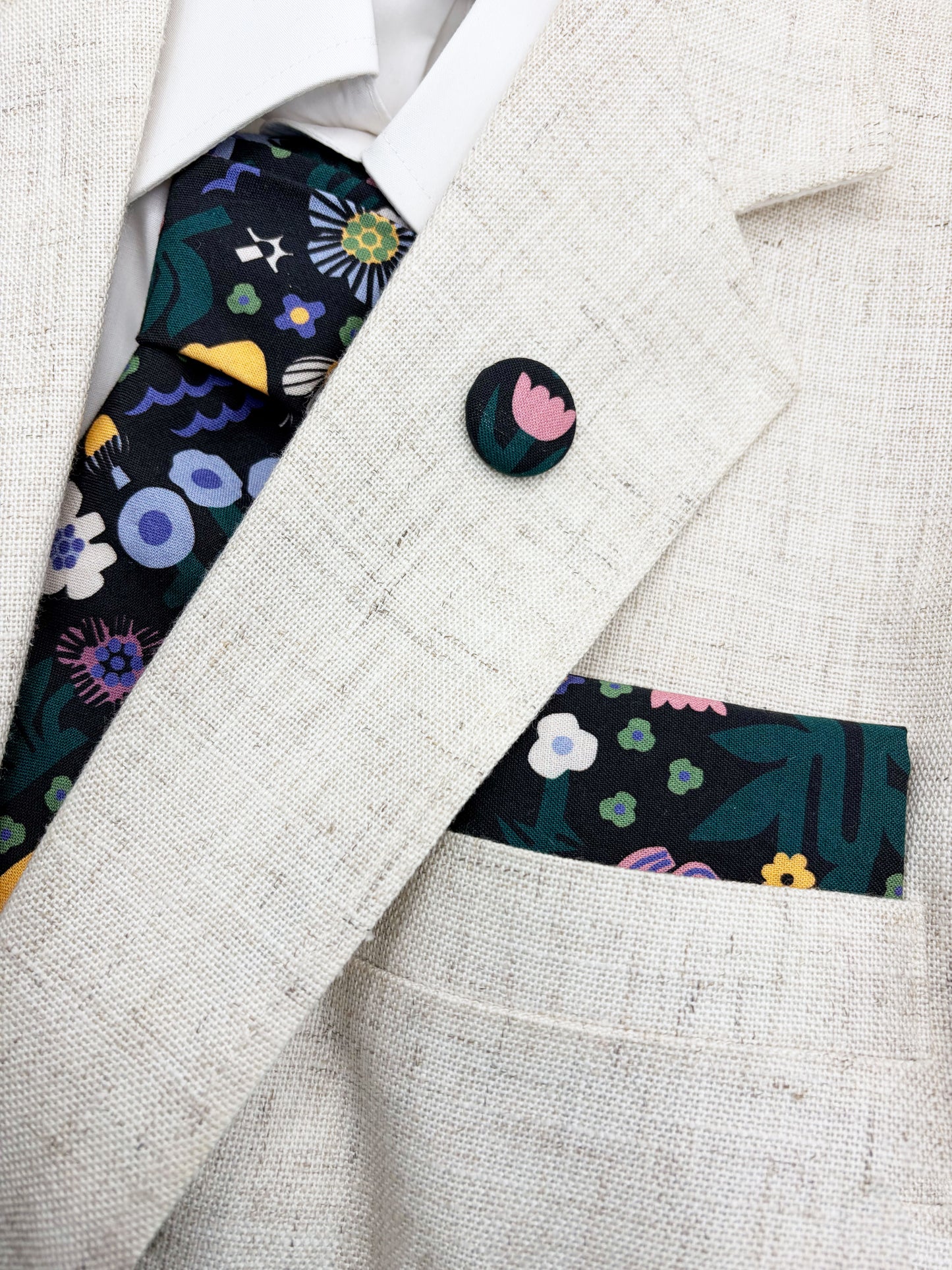 A necktie with an abstract print featuring bright forest creatures and flowers on a black background along with a matching pocket square and lapel pin.