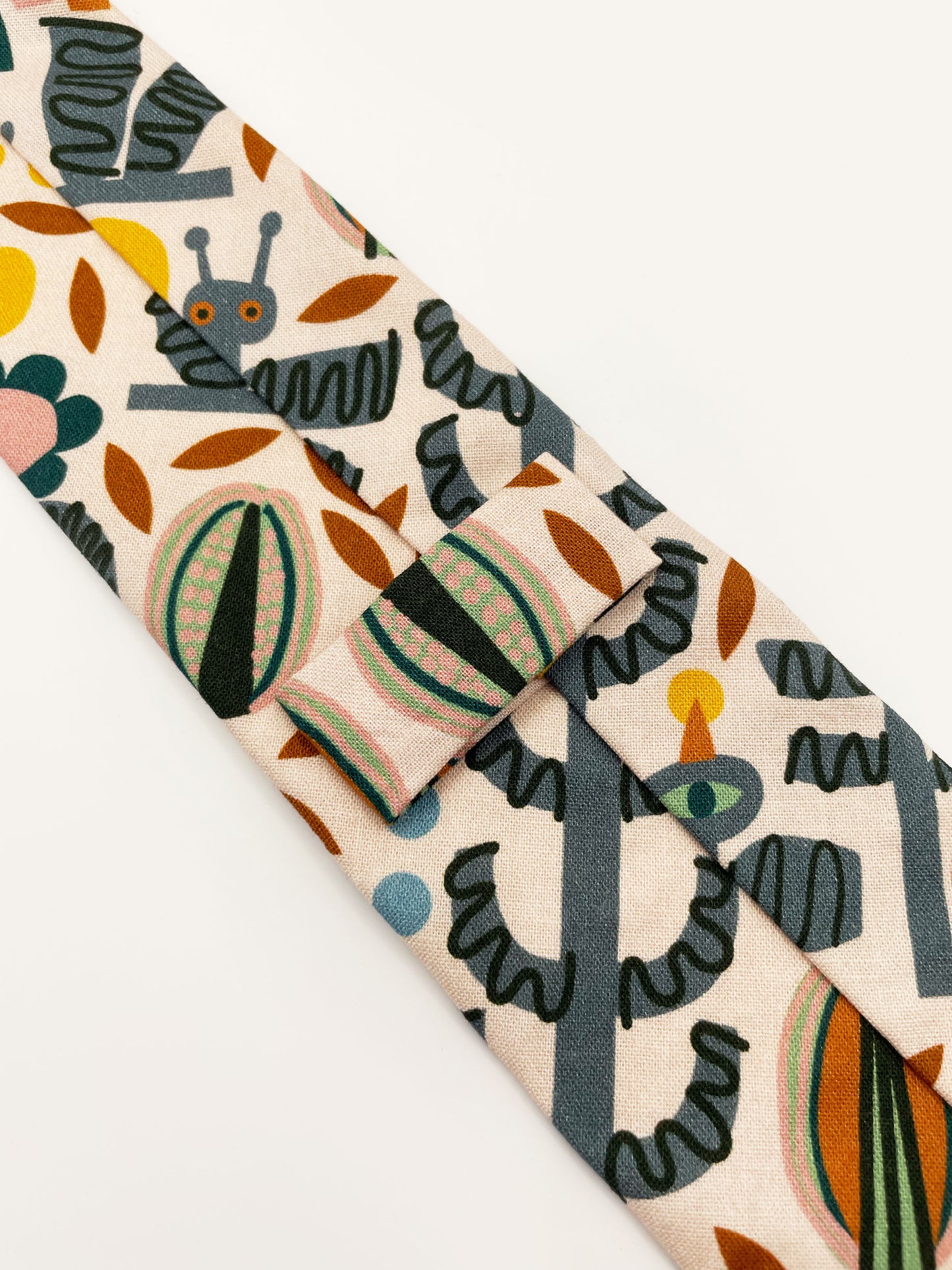 A necktie featuring a bold nature-inspired print of bugs, plants, suns, and rainbows. The color palette includes muted mustard, gentle gray, and soft pastels.
