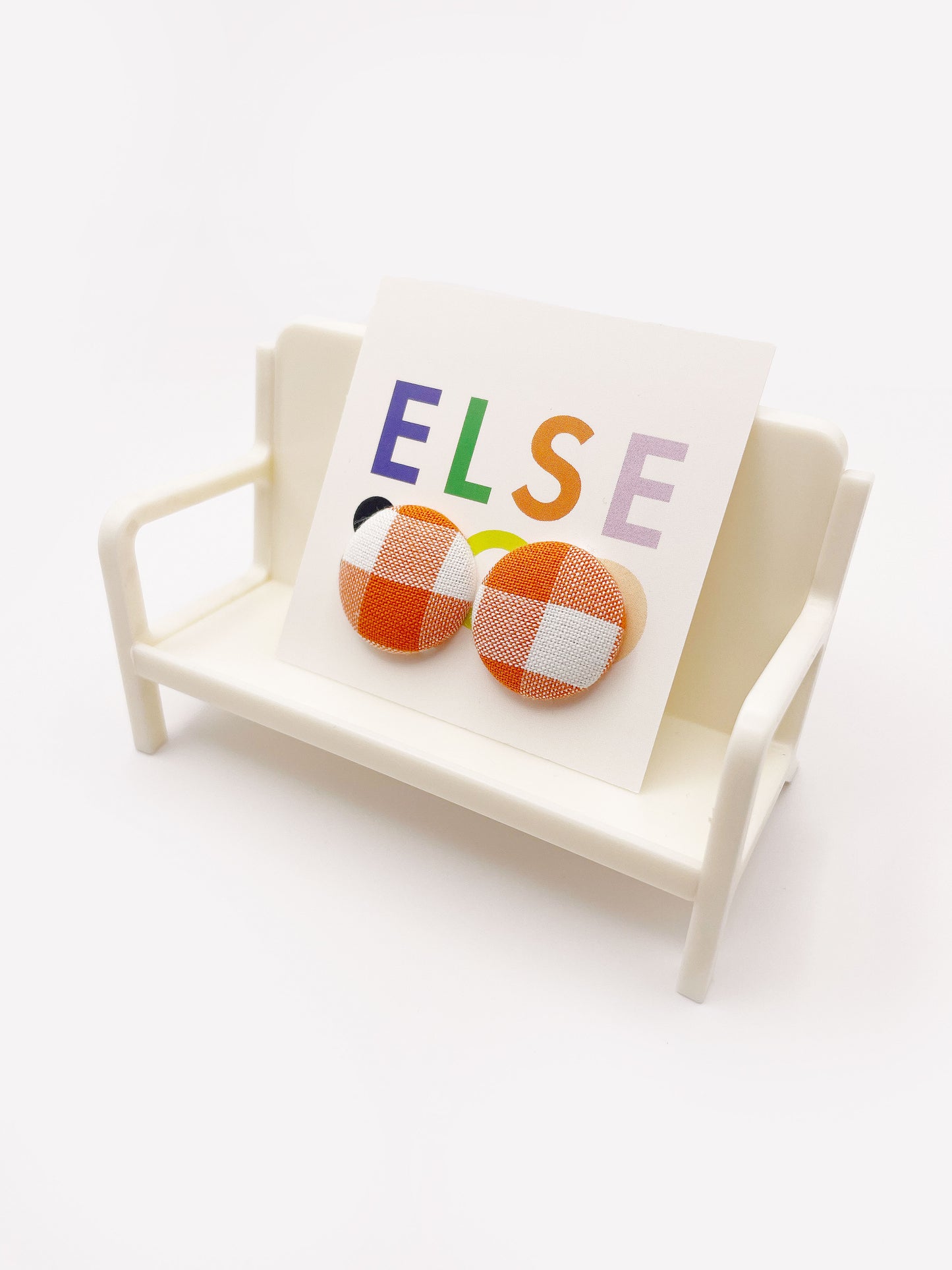 Handmade orange gingham plaid fabric covered post earrings. The earrings are on a backing card that says "Else Co". The earrings are shown on a small white plastic chair.