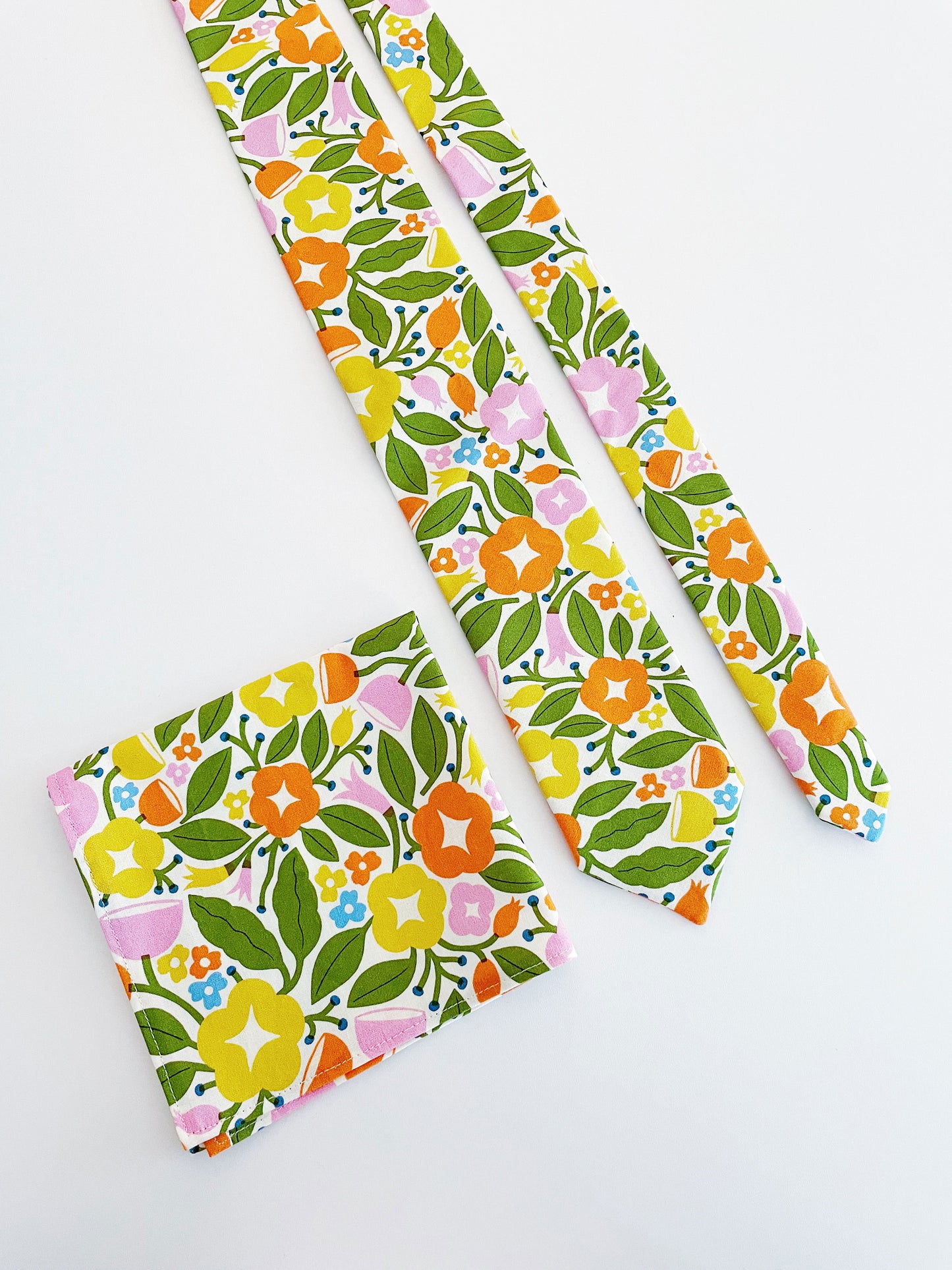 A vibrant pocket square with bold yellow, orange, and pink flowers. The pocket square is next to a matching necktie. The necktie is sold separately.