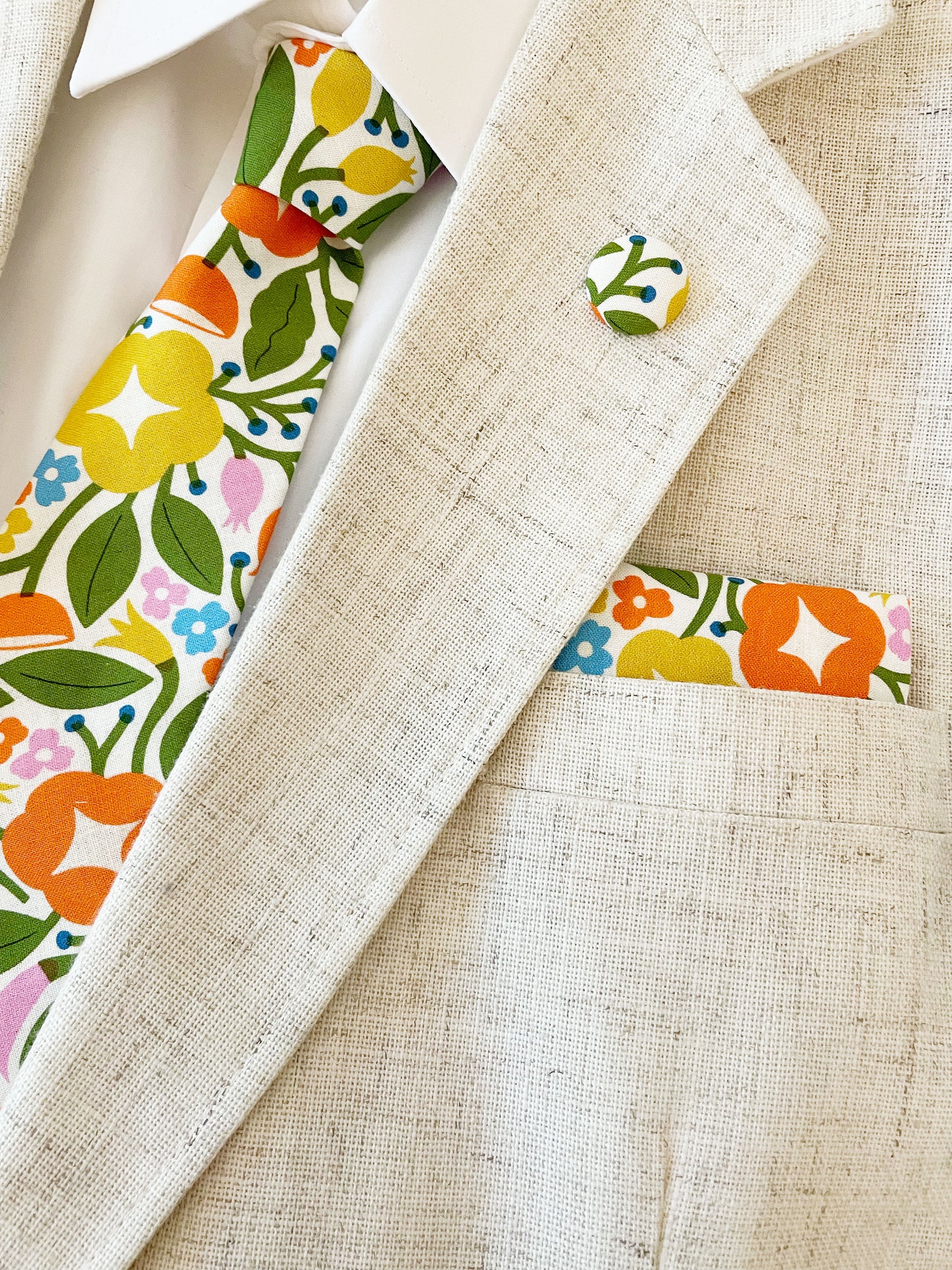 A vibrant necktie with bold yellow, orange, and pink flowers. The necktie is modeled with a white shirt and light tan jacket along with a matching pocket square and lapel pin. The pocket square is sold separately.