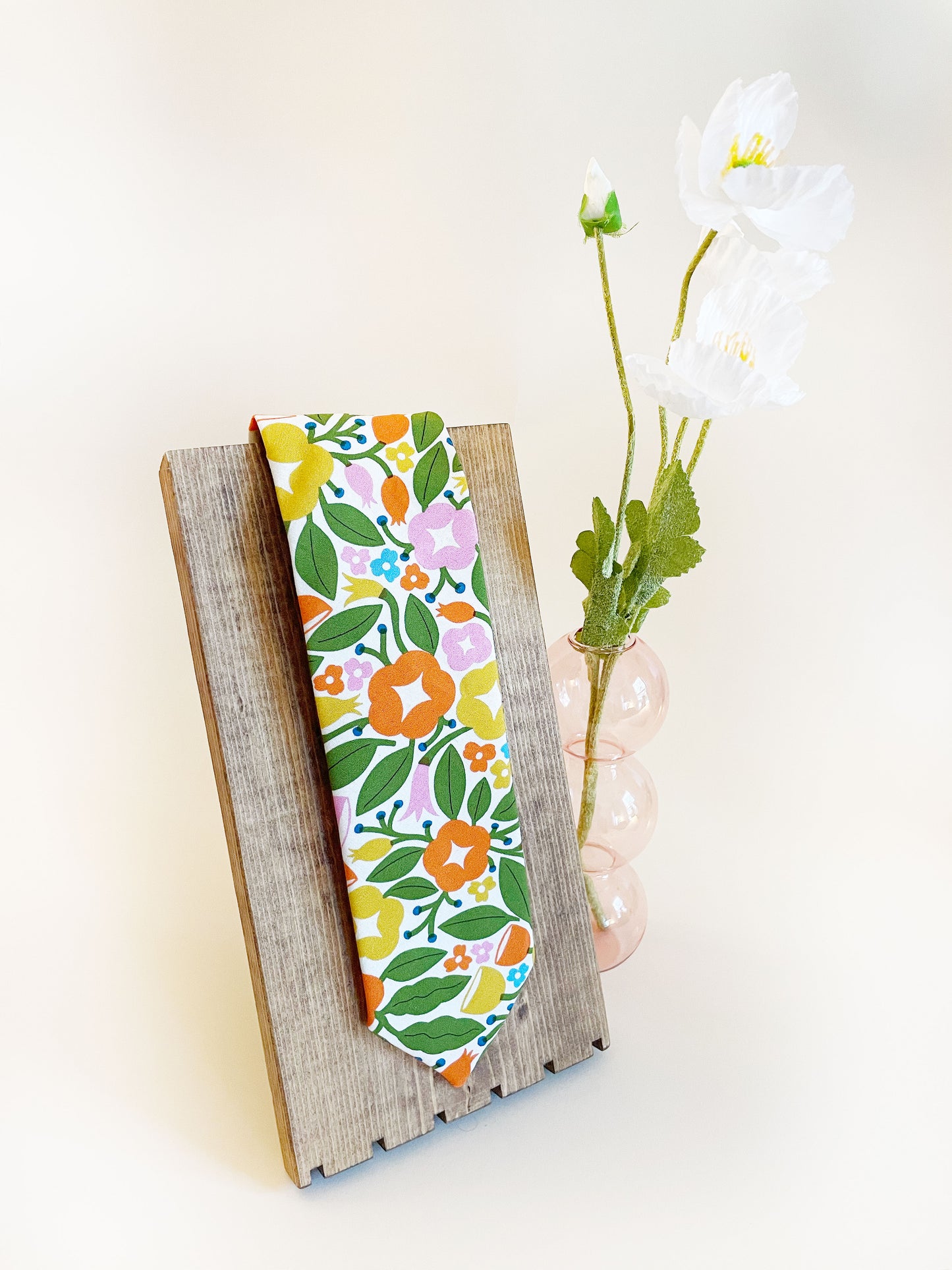 A vibrant necktie with bold yellow, orange, and pink flowers. The necktie is displayed on a wooden necklace stand next to a pink bubble vase containing white artificial flowers.