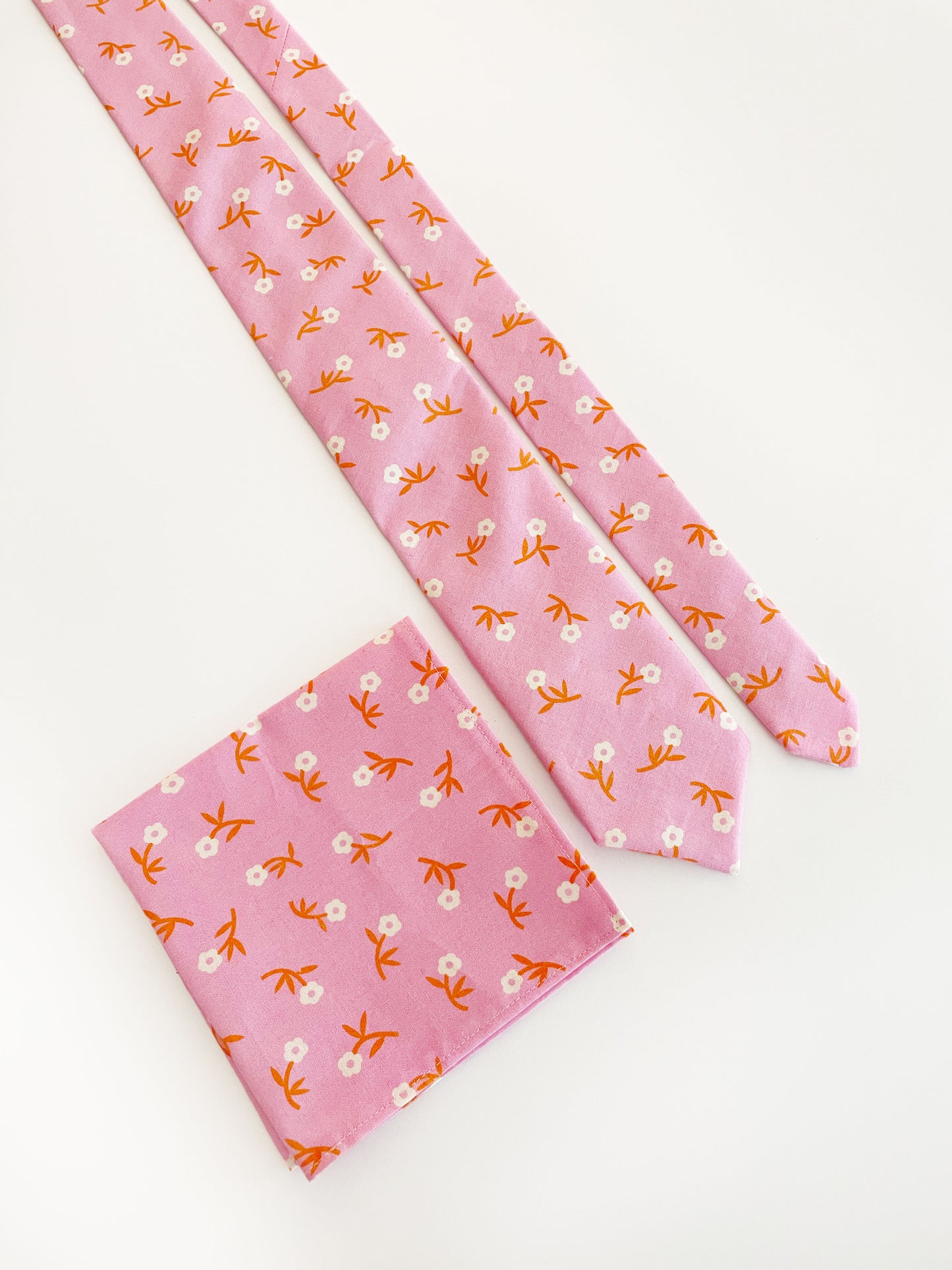 A cheerful pink pocket square with orange-stemmed flowers. The pocket square is next to a matching necktie. The necktie is sold separately.