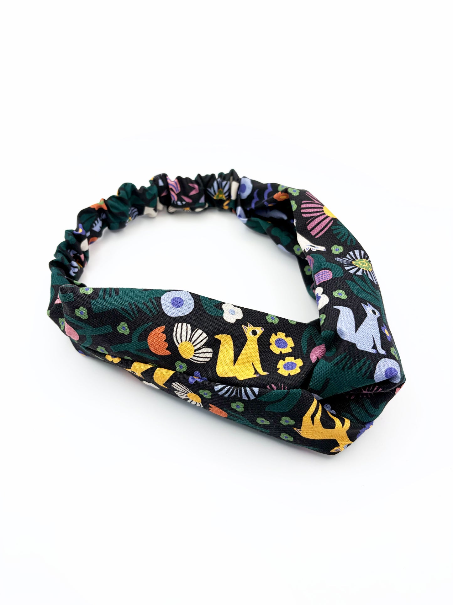 A black twist headband with a colorful abstract forest print featuring playful florals, leaves, and bright animals. The headband has an elastic band on the back.