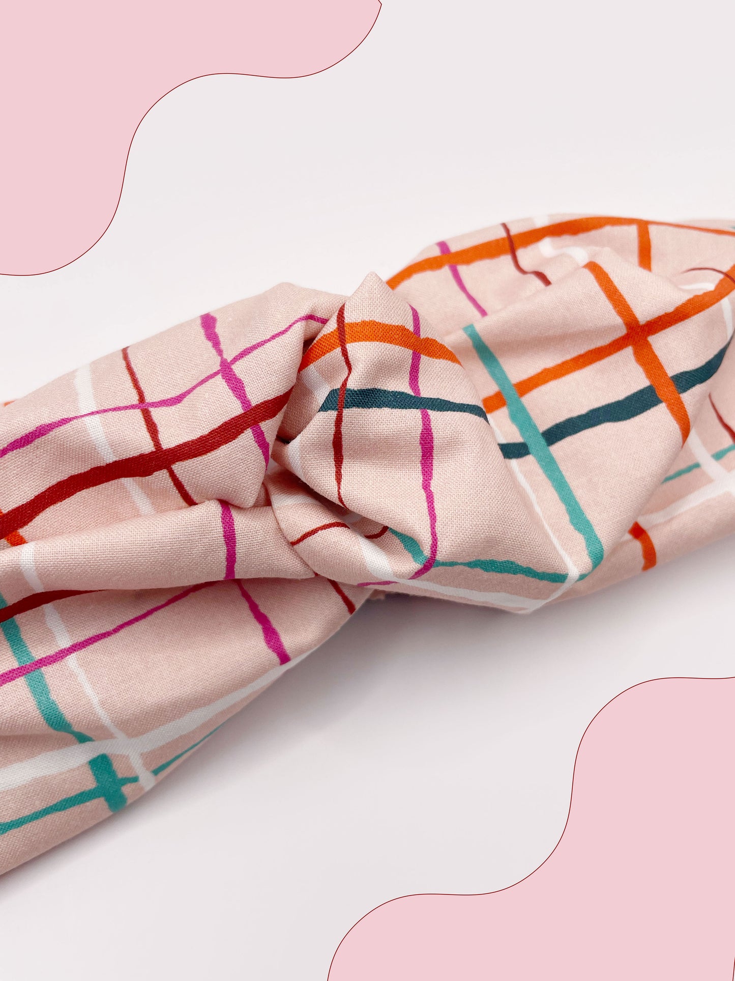 A fabric headband featuring a colorful and modern plaid design with vibrant shades of pink, red, teal, and orange on a soft pastel background.