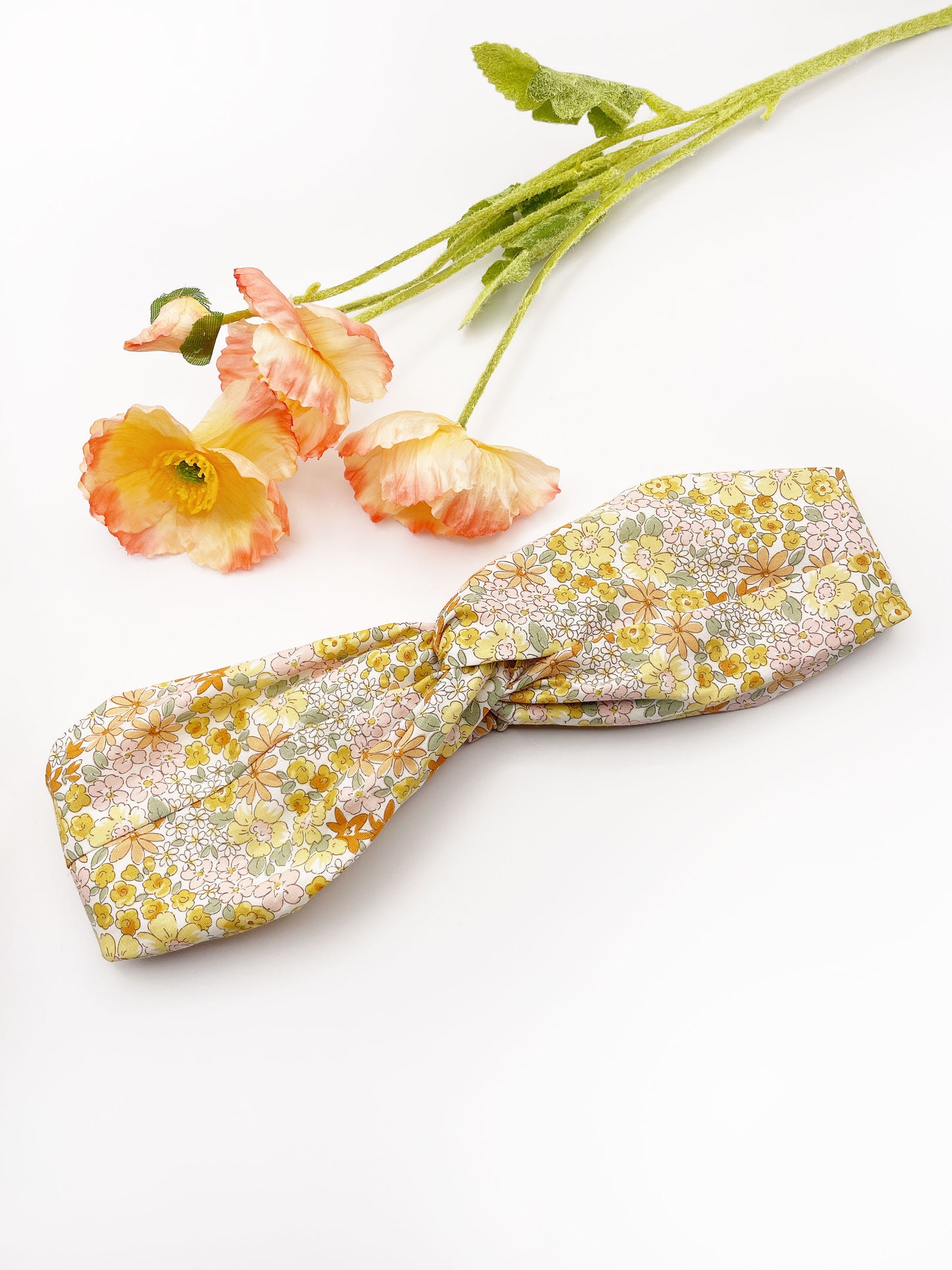 A handmade yellow floral twist headband with an elastic band in the back. The headband is next to artificial flowers.