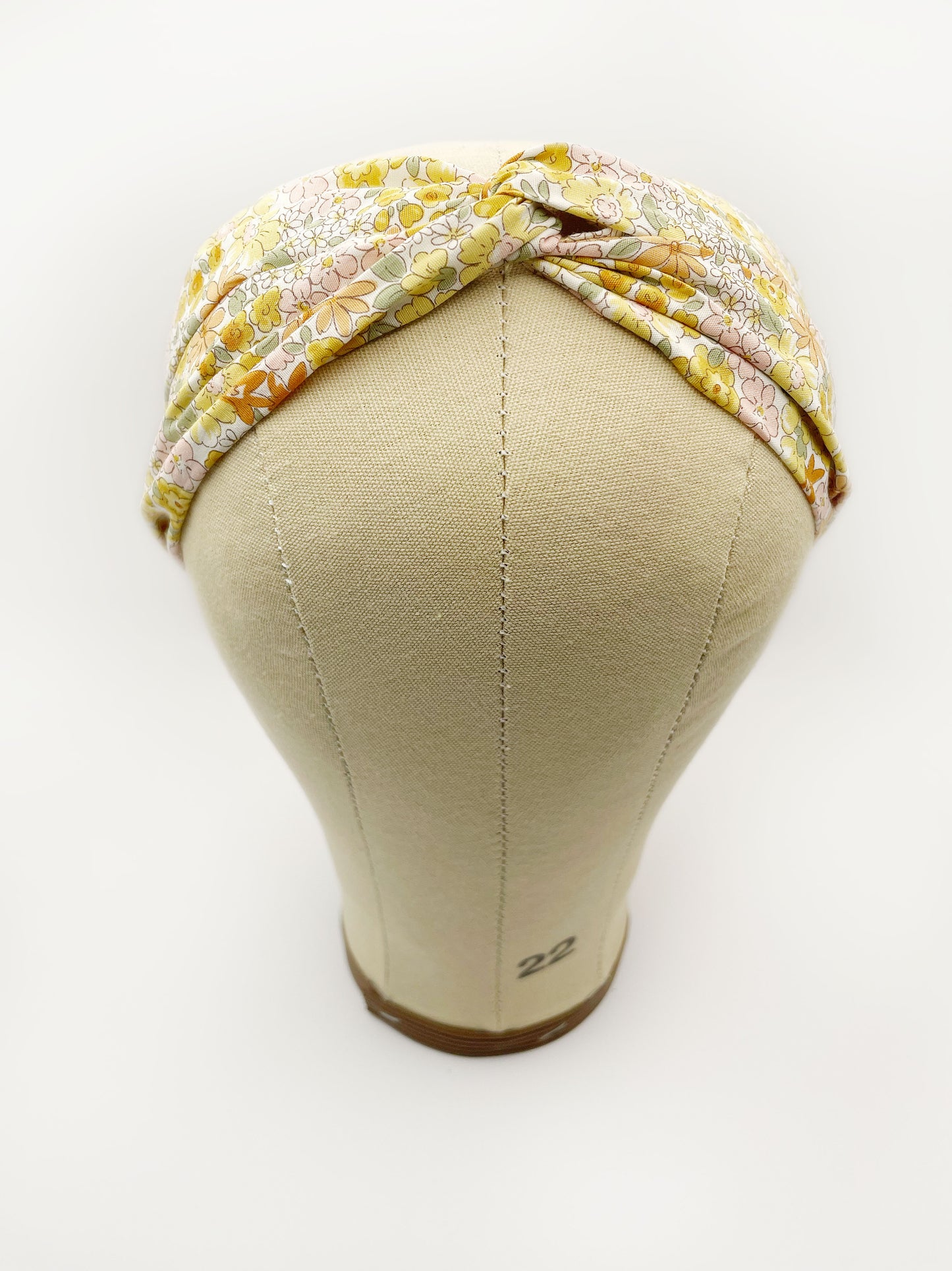 A handmade yellow floral twist headband with an elastic band in the back. The headband is modeled on a mannequin head.