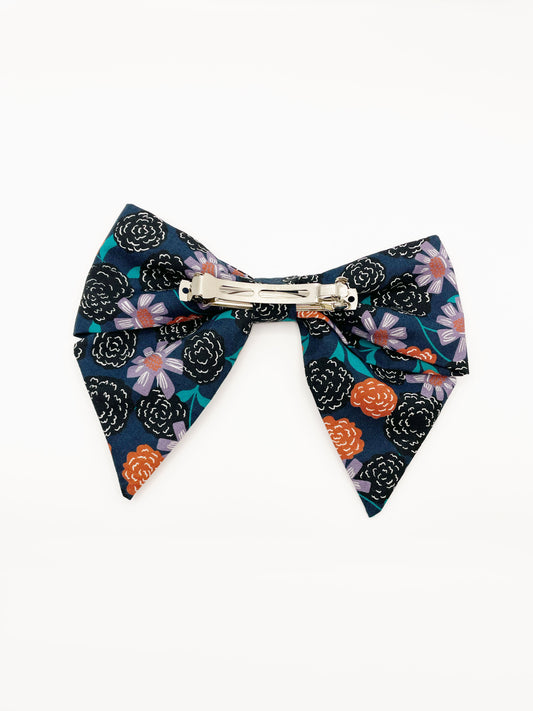 A large handmade hair bow with vivid orange, lavender, and black flowers, accented with fresh green leaves. The cotton fabric is attached to a sturdy barrette clip.