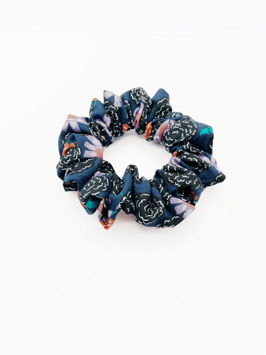 A scrunchie with vivid orange, lavender, and black flowers, accented with fresh green leaves.