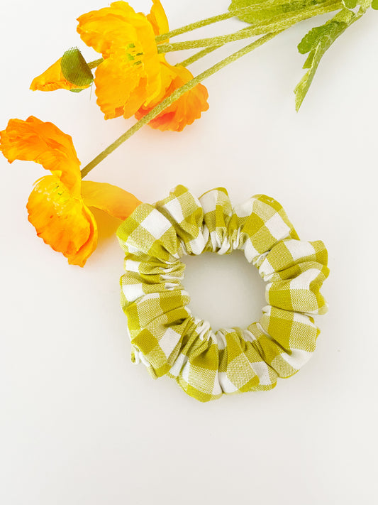 A green and white gingham plaid scrunchie. The scrunchie is next to orange artificial flowers.