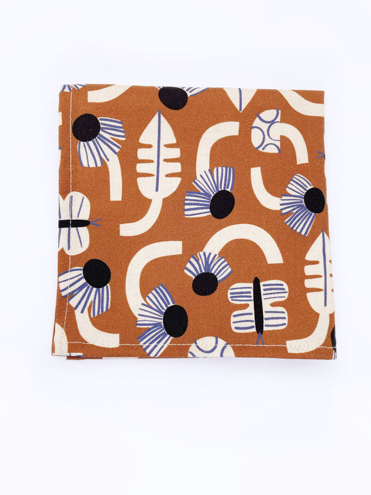 A pocket square with an abstract print featuring flowers, leaves, mushrooms, and butterflies on a rich terracotta background. The color palette also consists of cream, black, and blue.