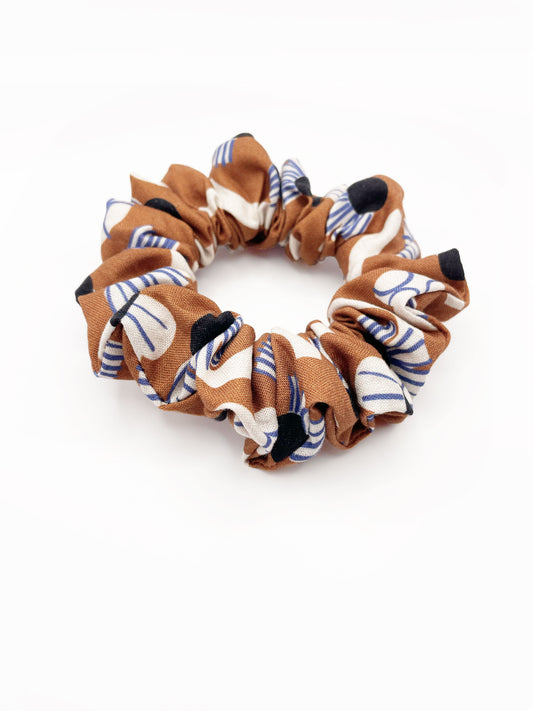 A scrunchie with an abstract print featuring flowers, leaves, and butterflies on a rich terracotta background. The color palette also consists of cream, black, and blue.