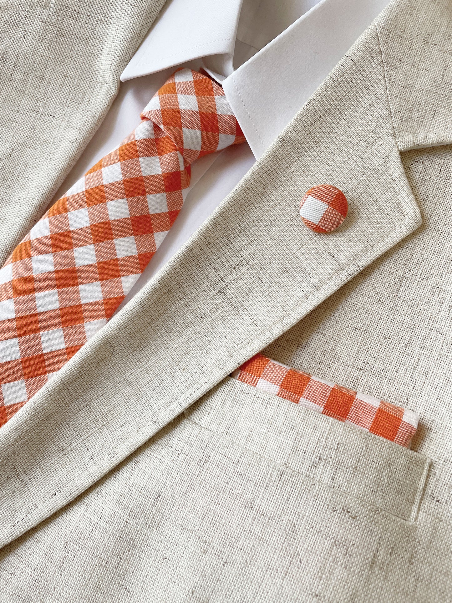 A cotton necktie with an orange and white gingham plaid print. The necktie is modeled with a matching pocket square and lapel pin.