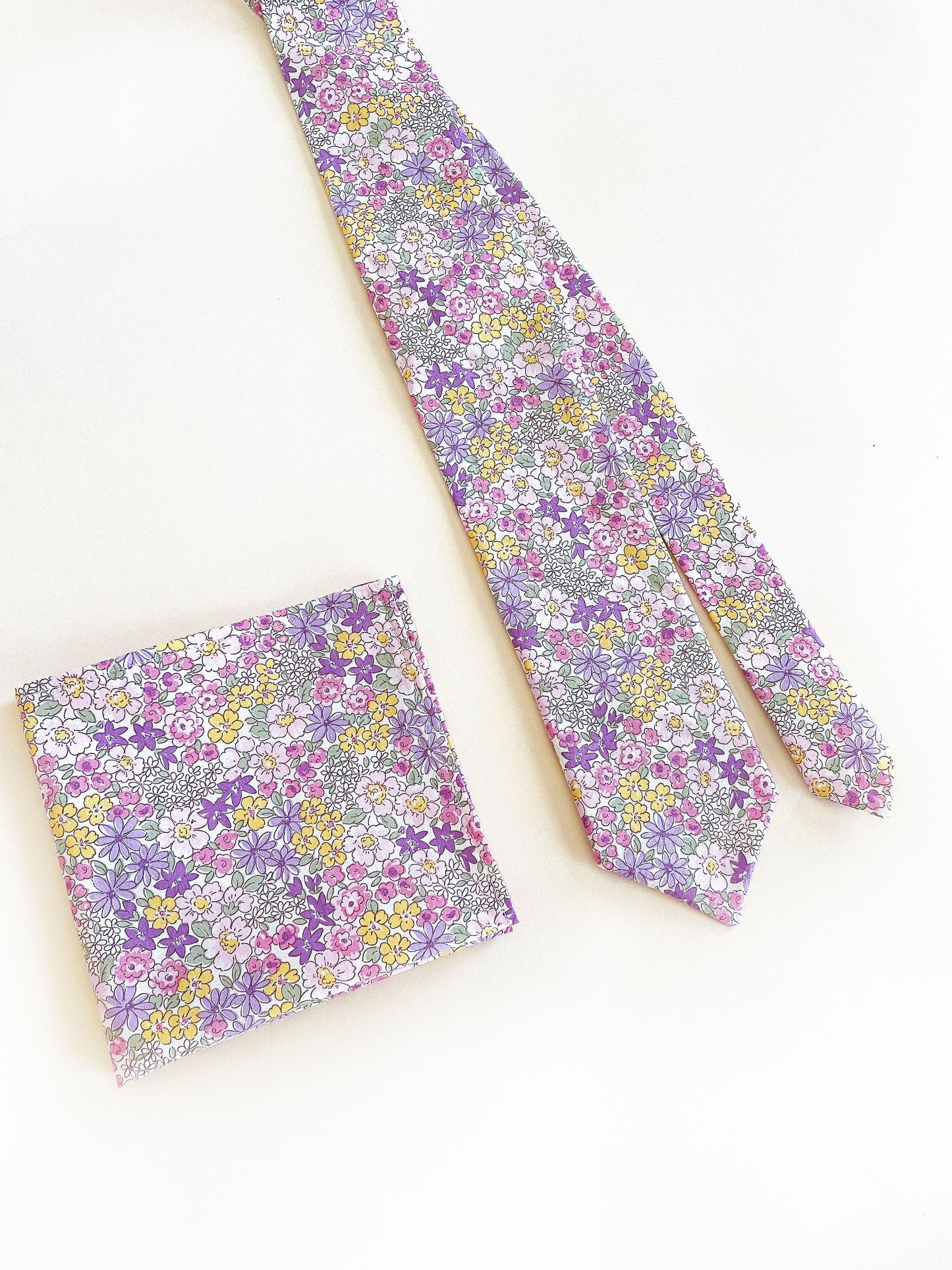A handmade purple floral necktie with a matching pocket square against an off white background.