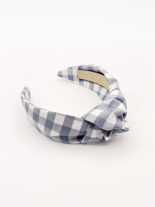 A handmade slate blue gingham plaid knotted headband against an off white background.