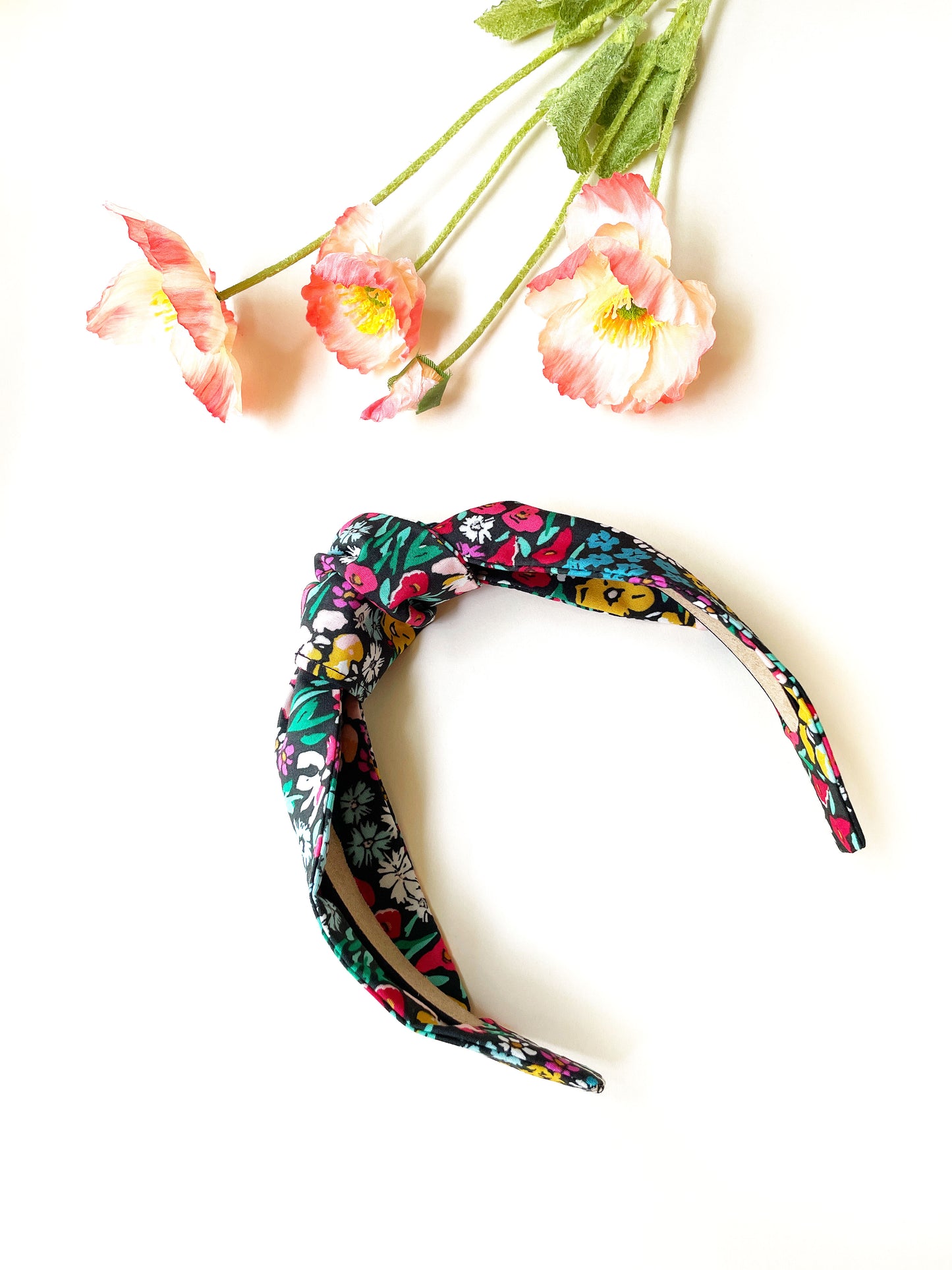 A black handmade knotted headband with a colorful floral print next to pink flowers.