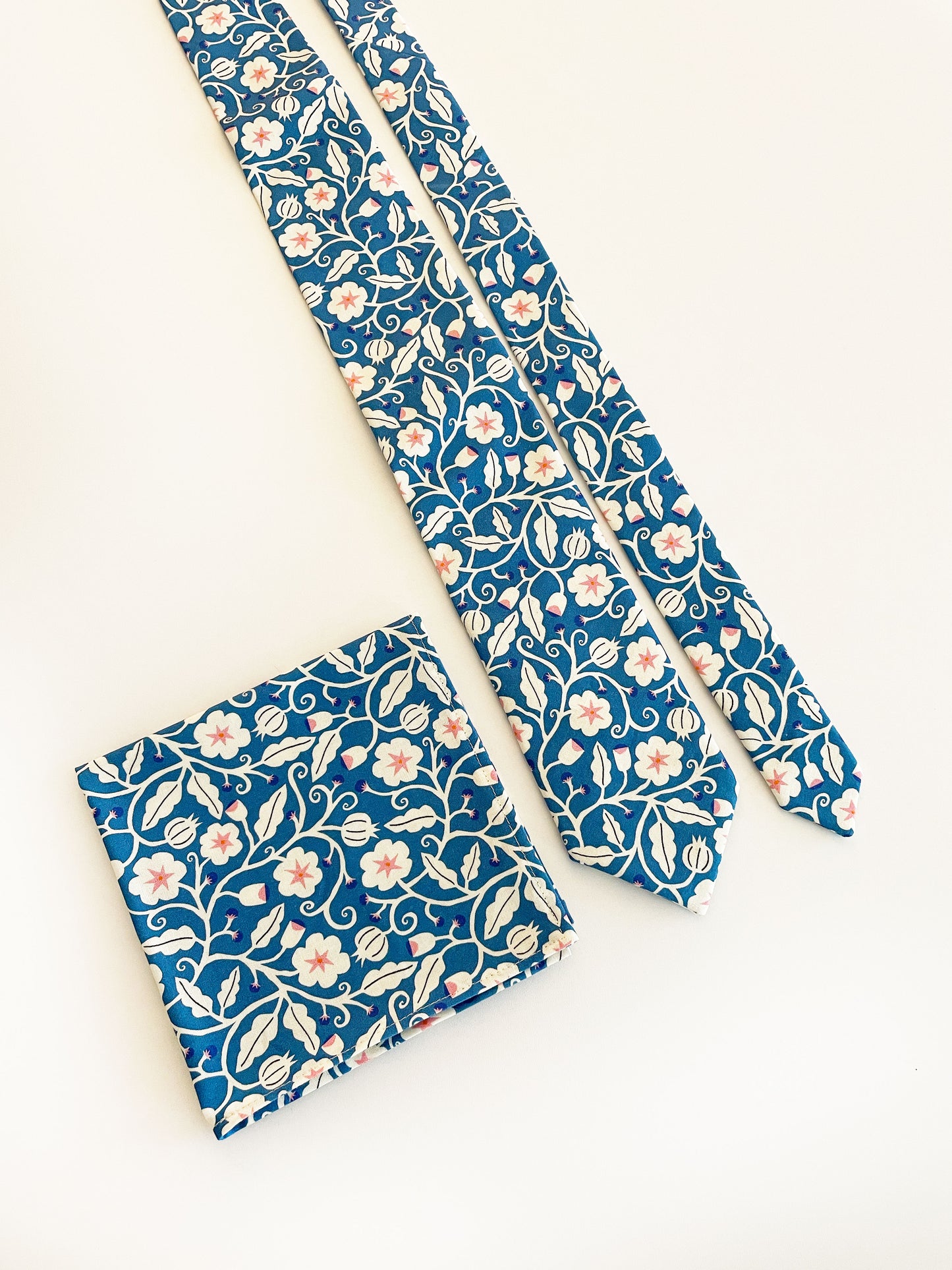 A blue pocket square with white flowers that have pink centers. The pocket square is next to a matching necktie. The necktie is sold separately.