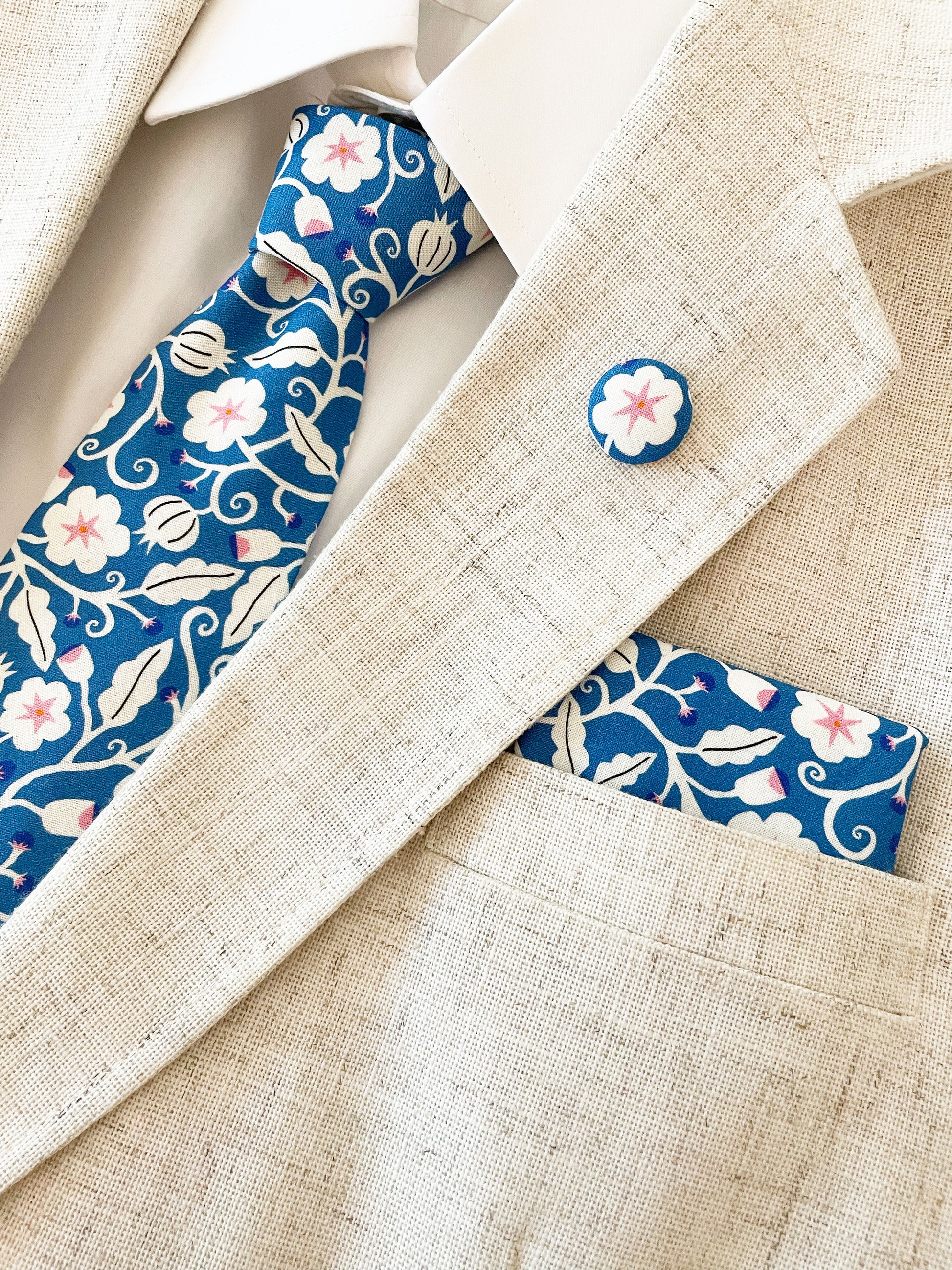 A blue pocket square with white flowers that have pink centers. The pocket square is modeled with a matching necktie and lapel pin. The necktie is sold separately.