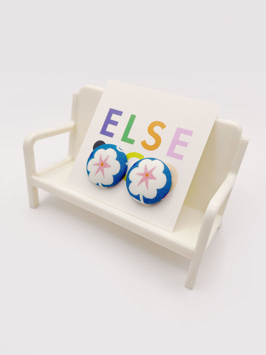 Handmade blue fabric-covered earrings with white flowers that have pink centers. The earrings are on a backing card that says "Else Co". The earrings are displayed on a small plastic chair.