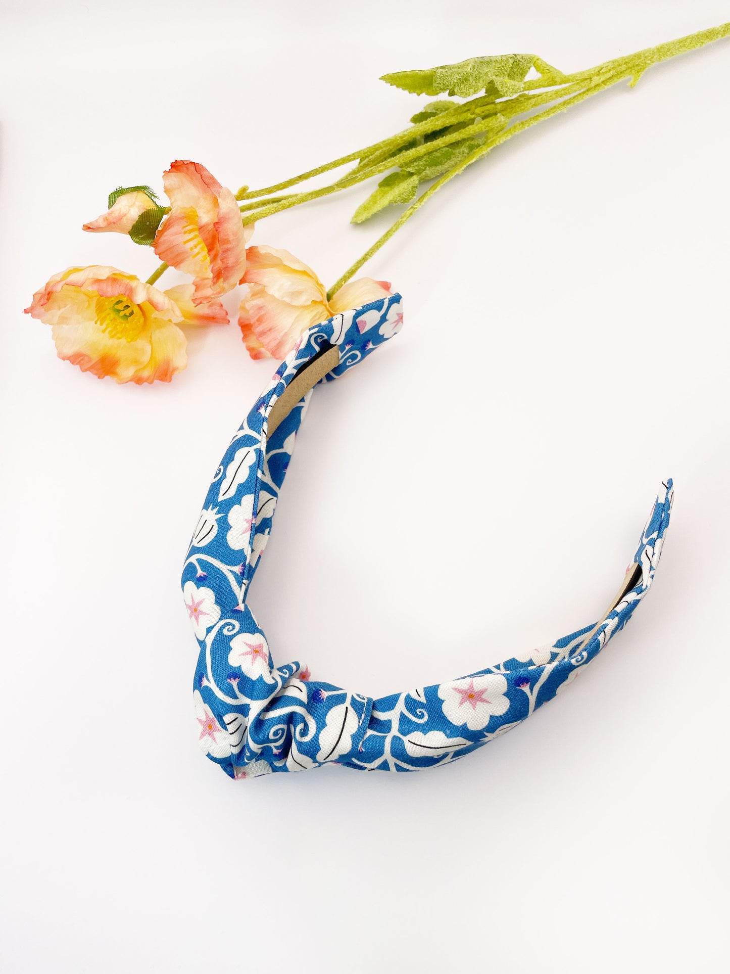 A blue knotted headband with white flowers that have pink centers. The headband is next to some pink artificial flowers.