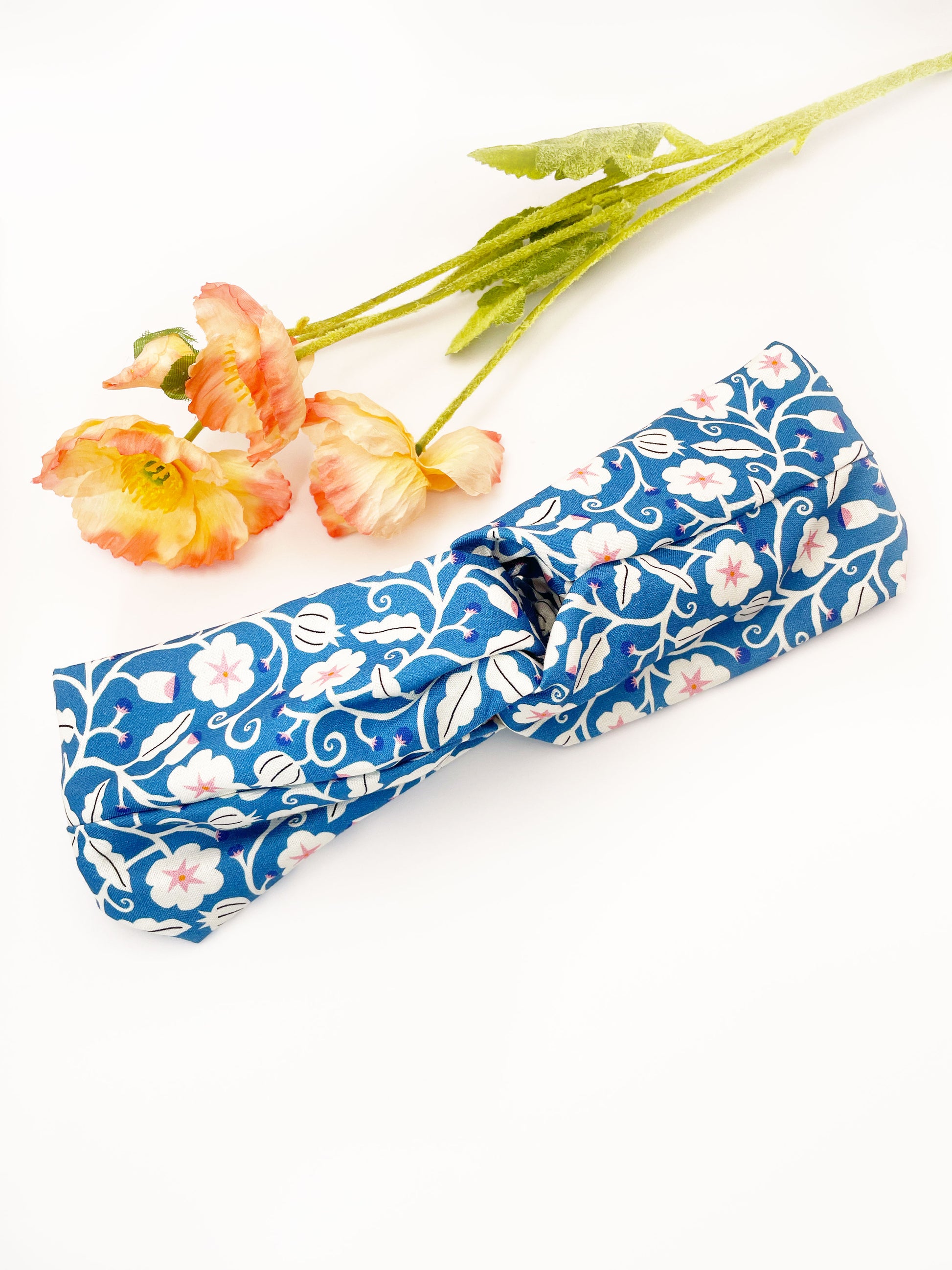 A blue twist headband with white flowers that have pink centers. The headband is next to some pink artificial flowers.