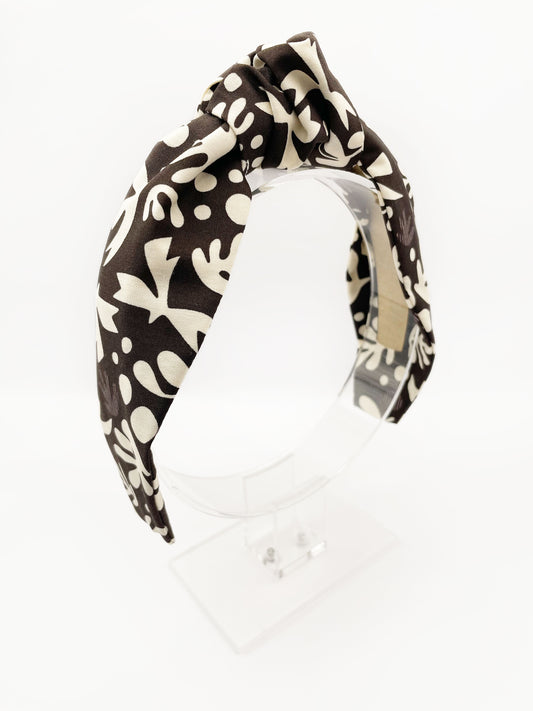 A dark charcoal gray knotted headband with cream-colored birds and abstract shapes. The headband is displayed on an acrylic stand.