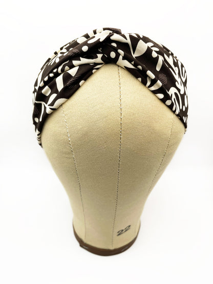 A dark charcoal gray twist headband with cream-colored birds and abstract shapes. The headband is modeled on a mannequin head.