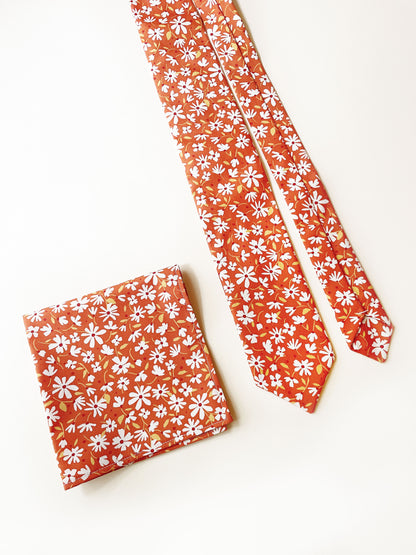 A rust brown floral pocket square with white flowers, showcasing shades of earthy browns and pops of warm peach and blush pink next to a matching necktie.