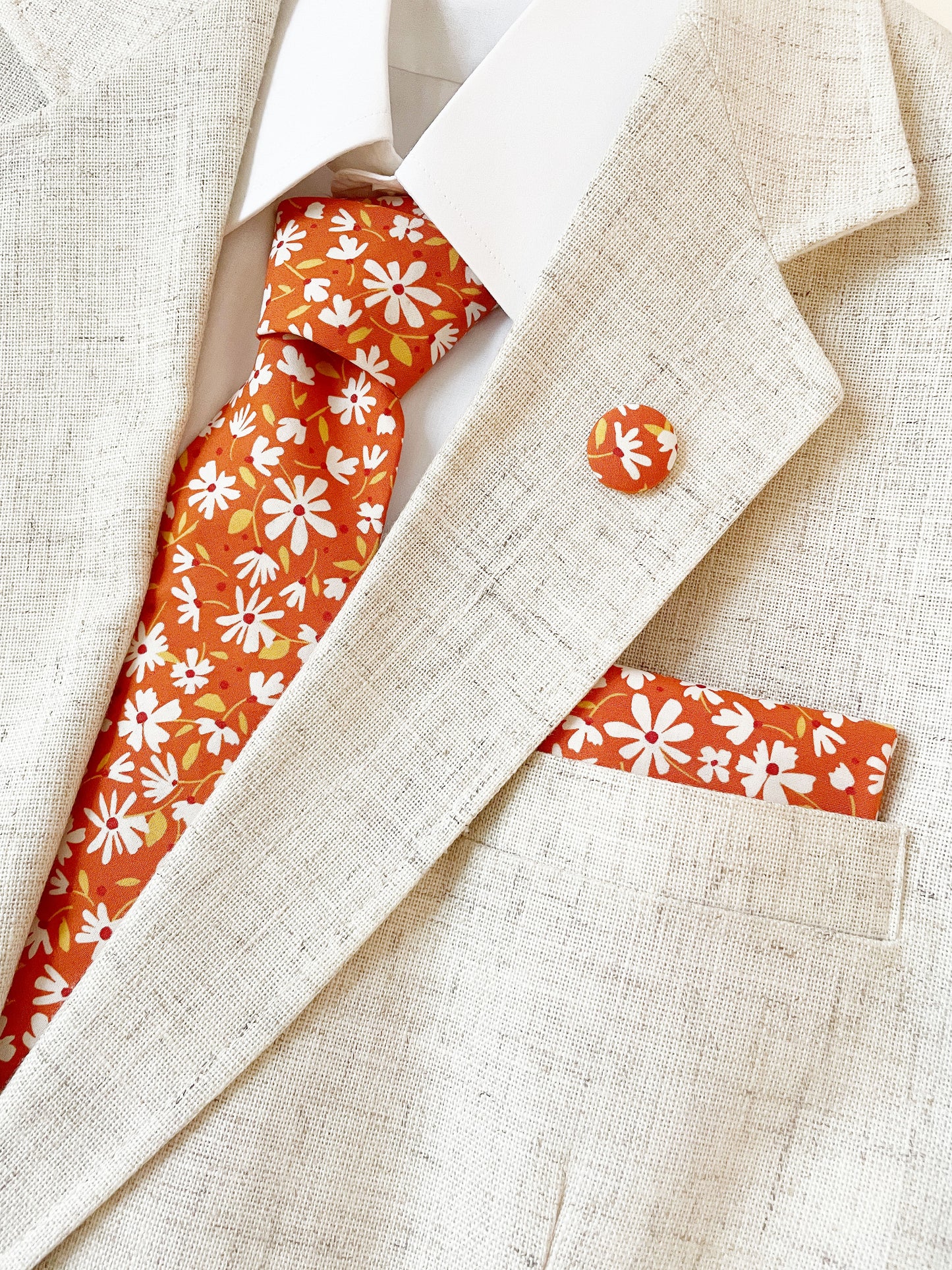 A rust brown floral pocket square with white flowers, showcasing shades of earthy browns and pops of warm peach and blush pink with a matching necktie and lapel pin.