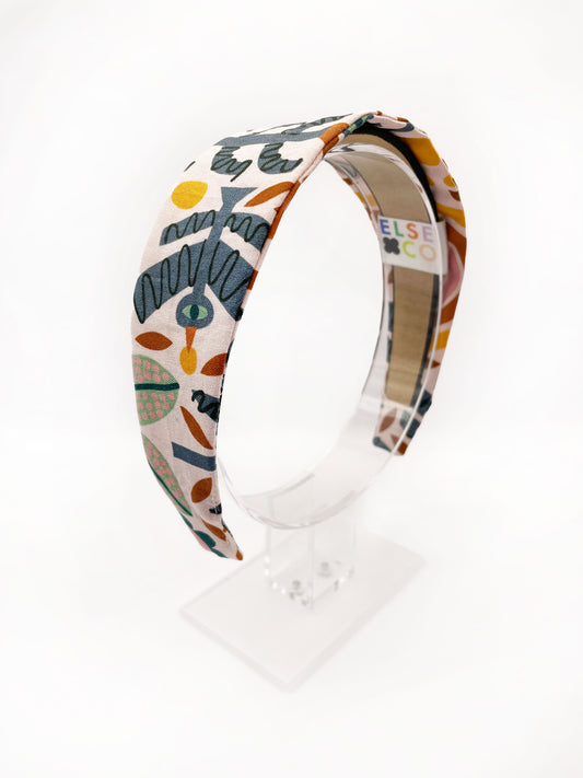 A fabric headband featuring a bold nature-inspired print of bugs, plants, suns, and rainbows. The headband is shown on an acrylic display.