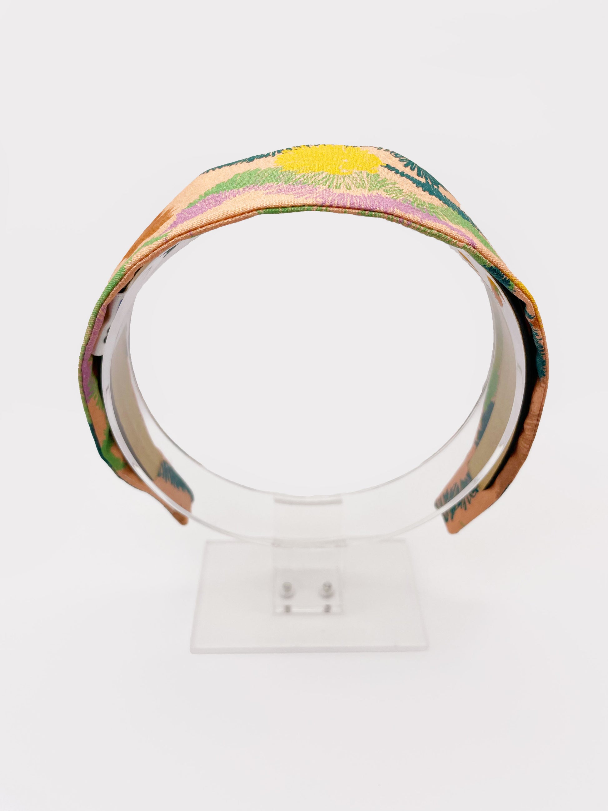 A fabric headband with an abstract design inspired by the desert. The color palette includes sandy brown, soft blue, muted green, and warm terracotta. The headband is shown on an acrylic display.