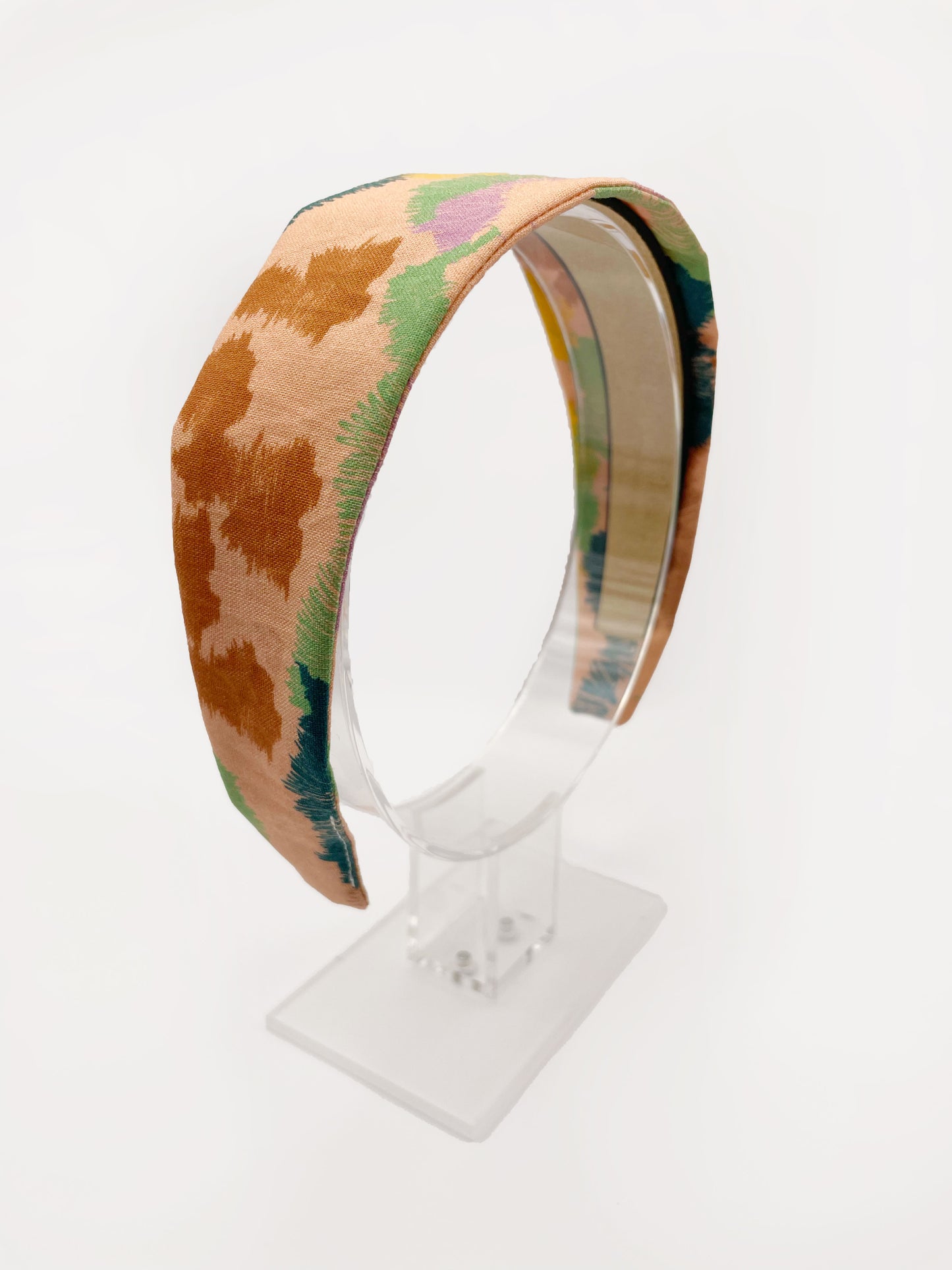 A fabric headband with an abstract design inspired by the desert. The color palette includes sandy brown, soft blue, muted green, and warm terracotta. The headband is shown on an acrylic display.