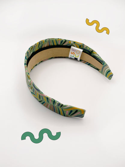 A fabric-covered headband featuring an abstract design inspired by desert rain patterns. The material is green with a soft brushstroke-like print, and a harmonious blend of yellow, pink, and blue.