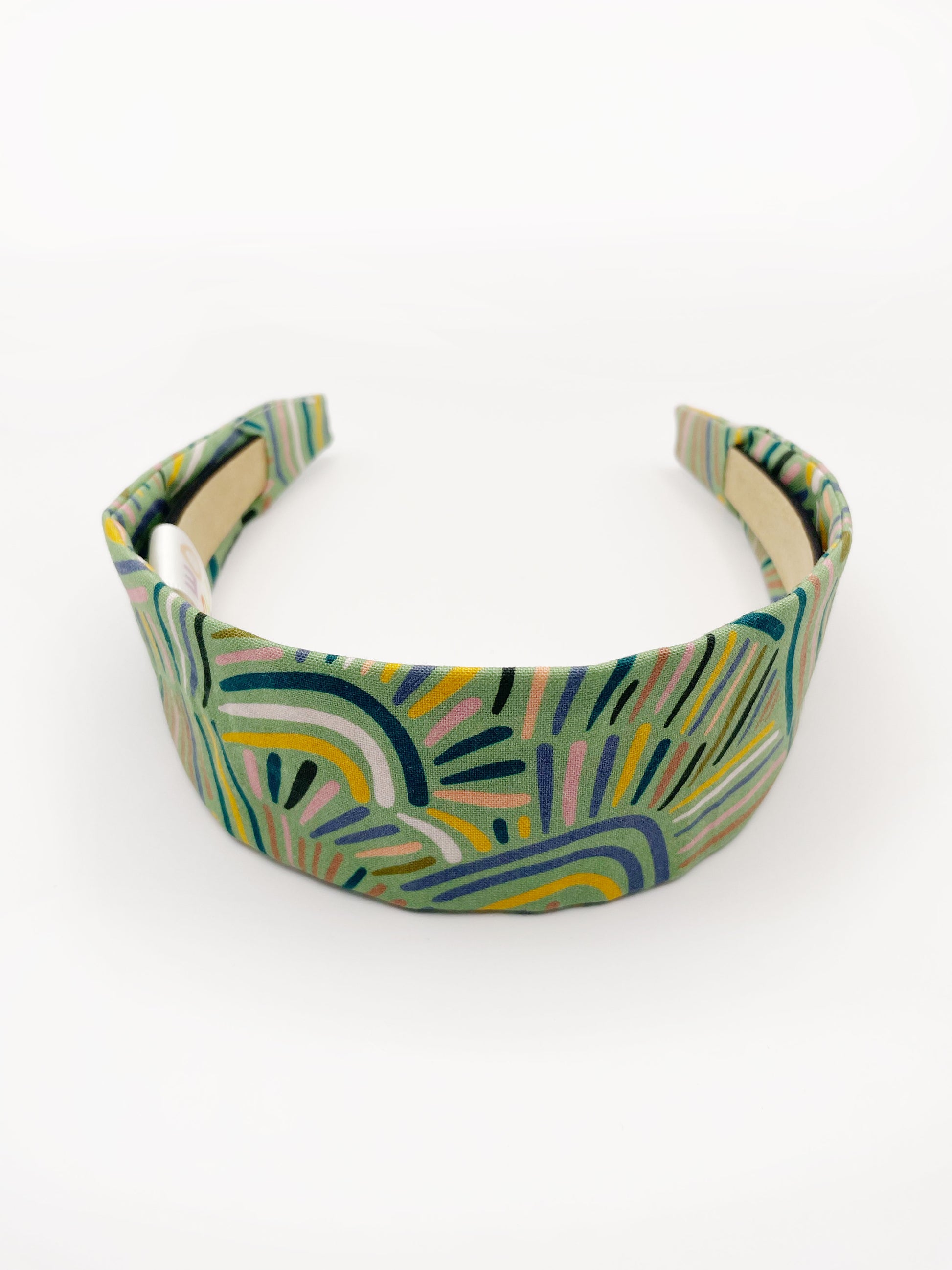 A fabric-covered headband featuring an abstract design inspired by desert rain patterns. The material is green with a soft brushstroke-like print, and a harmonious blend of yellow, pink, and blue.