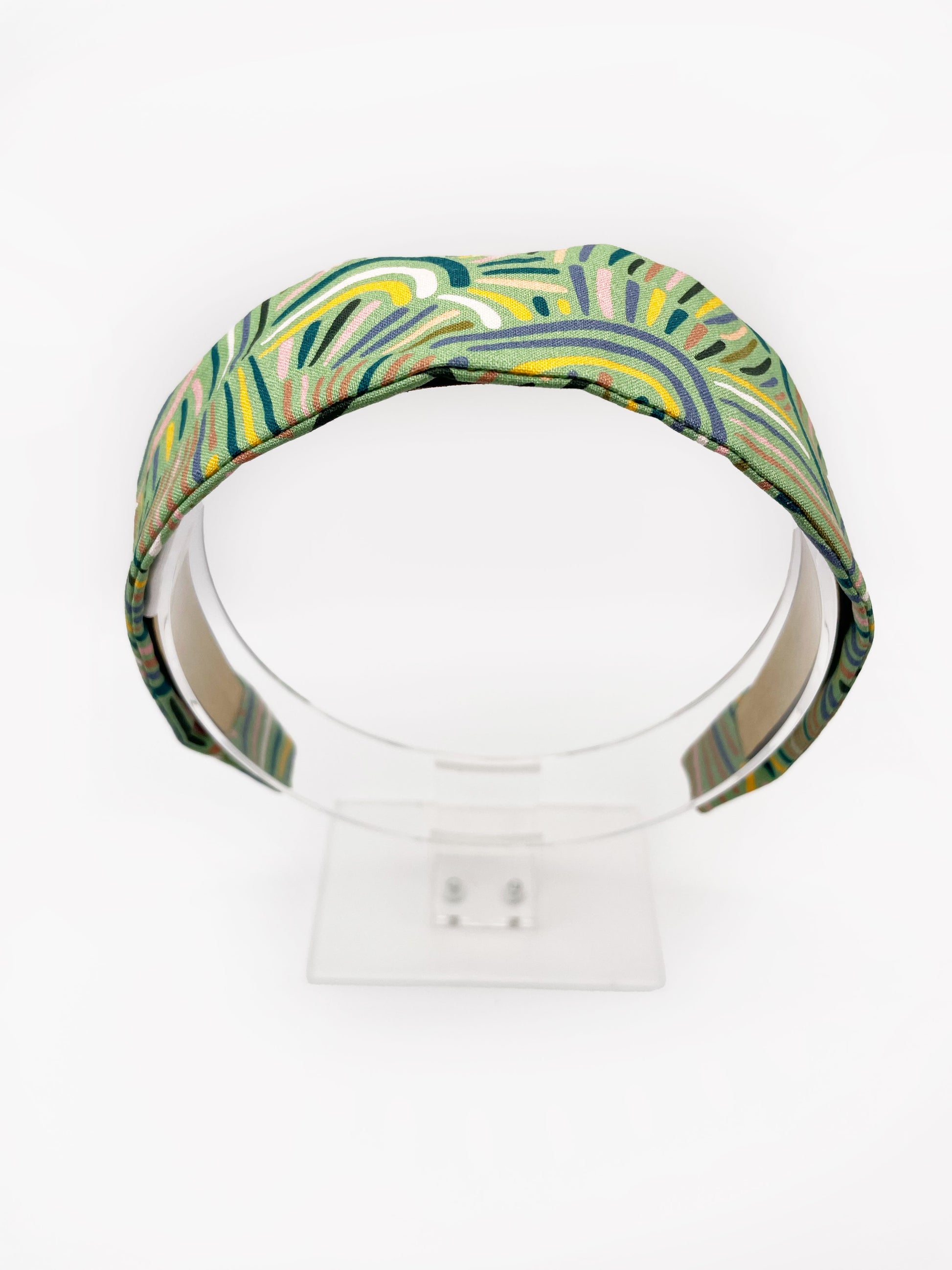 A fabric-covered headband featuring an abstract design inspired by desert rain patterns. The material is green with a soft brushstroke-like print, and a harmonious blend of yellow, pink, and blue. The headband is shown on an acrylic display.