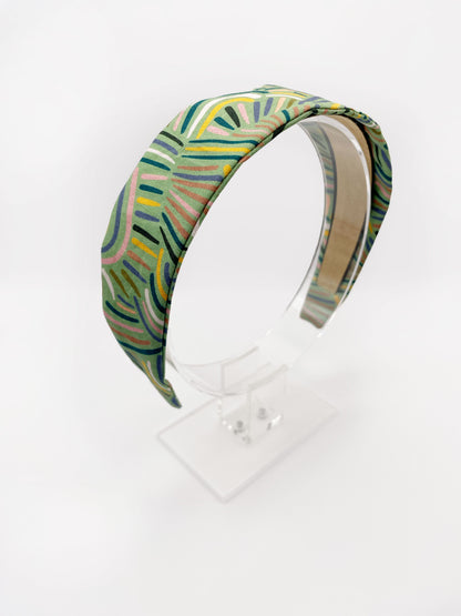 A fabric-covered headband featuring an abstract design inspired by desert rain patterns. The material is green with a soft brushstroke-like print, and a harmonious blend of yellow, pink, and blue. The headband is shown on an acrylic display.