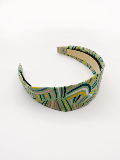 A fabric-covered headband featuring an abstract design inspired by desert rain patterns. The material is green with a soft brushstroke-like print, and a harmonious blend of yellow, pink, and blue.