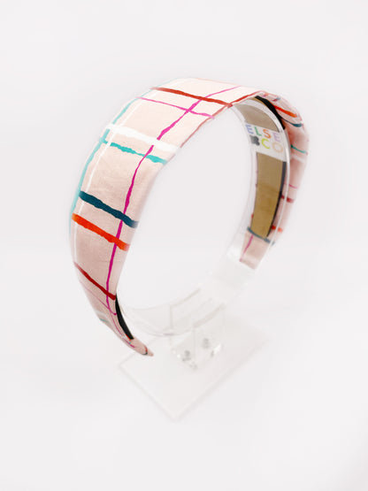 A fabric headband featuring a colorful and modern plaid design with vibrant shades of pink, red, teal, and orange on a soft pastel background. The headband is shown on an acrylic stand.