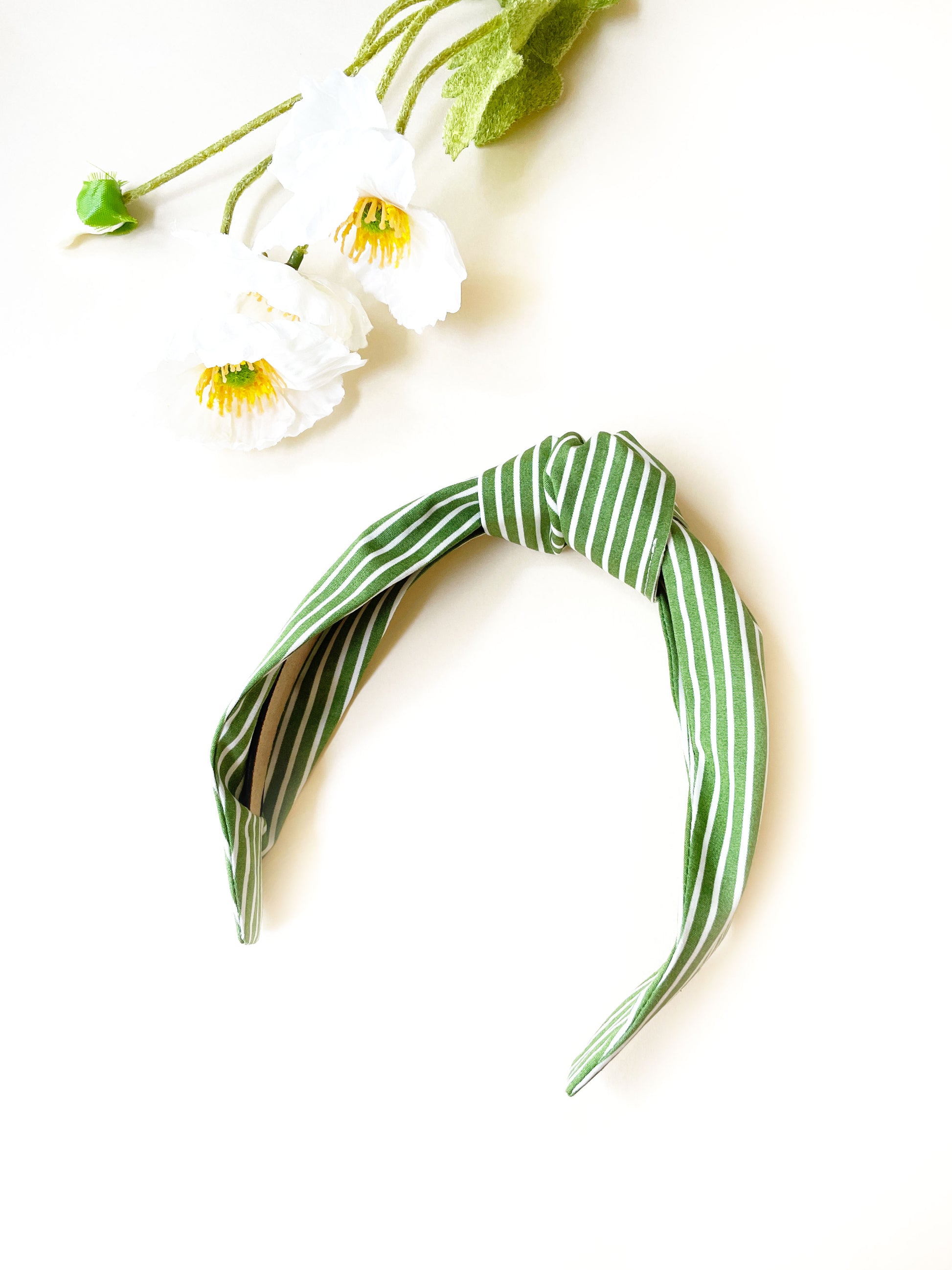 A handmade knotted headband featuring a white striped print against a moss green background next to some white flowers.