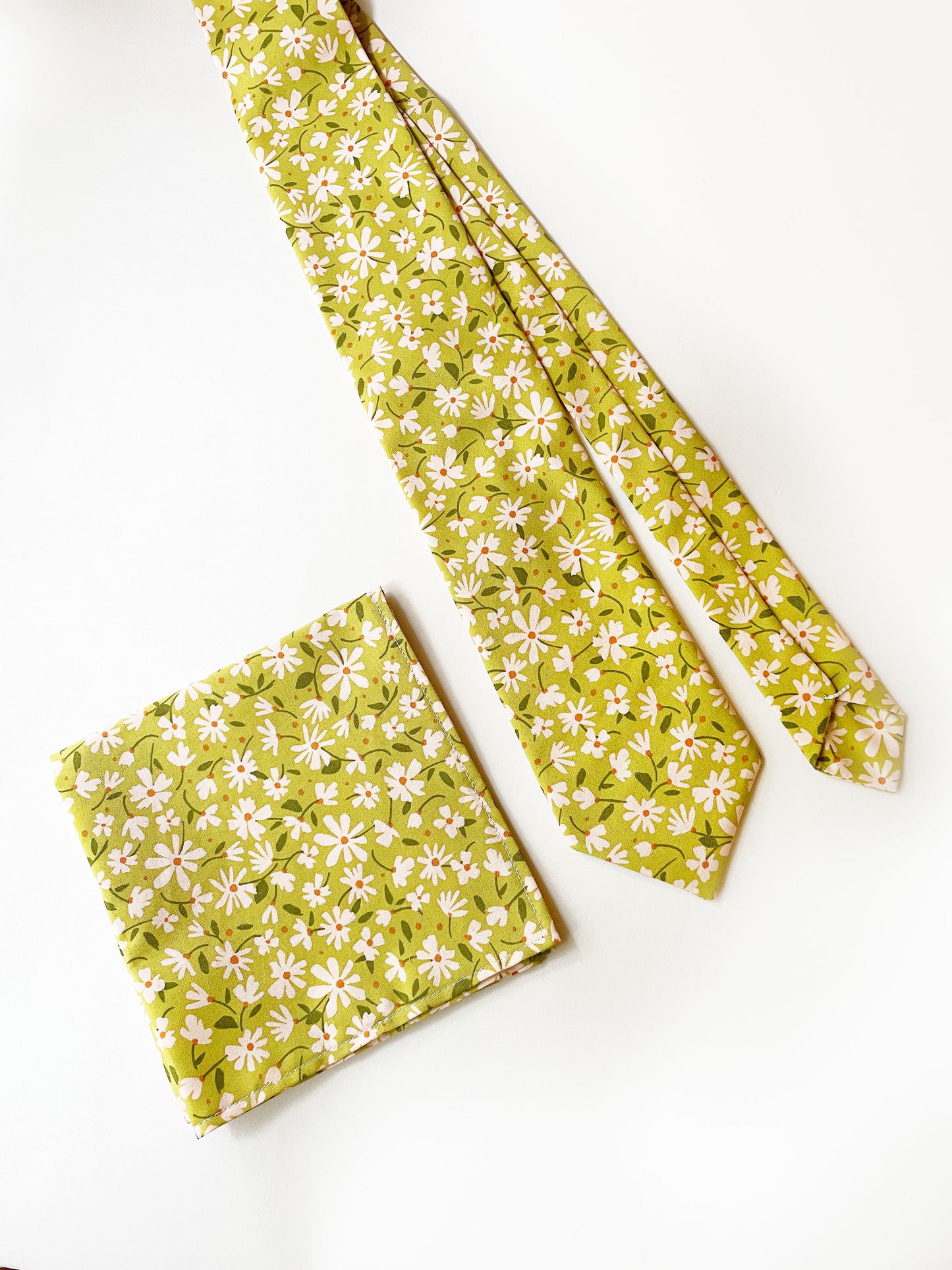 A mossy green handmade necktie with cream flowers, showcasing shades of earthy browns and pops of warm peach and blush pink. The tie is next to a matching pocket square.