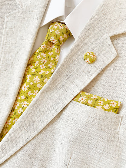 A mossy green floral pocket square with cream flowers, showcasing shades of earthy browns and pops of warm peach and blush pink with a matching necktie and lapel pin.