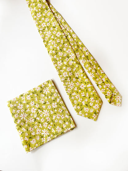 A mossy green floral pocket square with cream flowers, showcasing shades of earthy browns and pops of warm peach and blush pink next to a matching necktie.