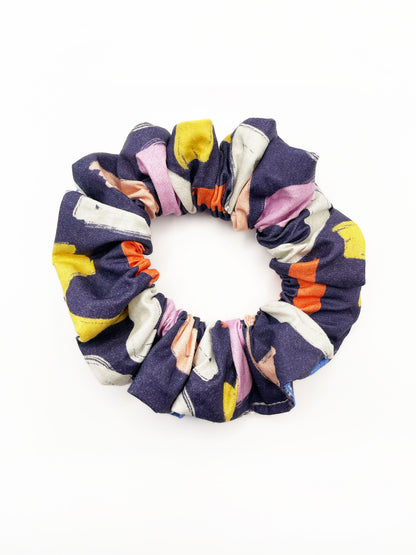 A navy blue scrunchie with a colorful abstract print featuring various doodles. Wear this hair tie on your wrist, or use it as a bun or ponytail holder.