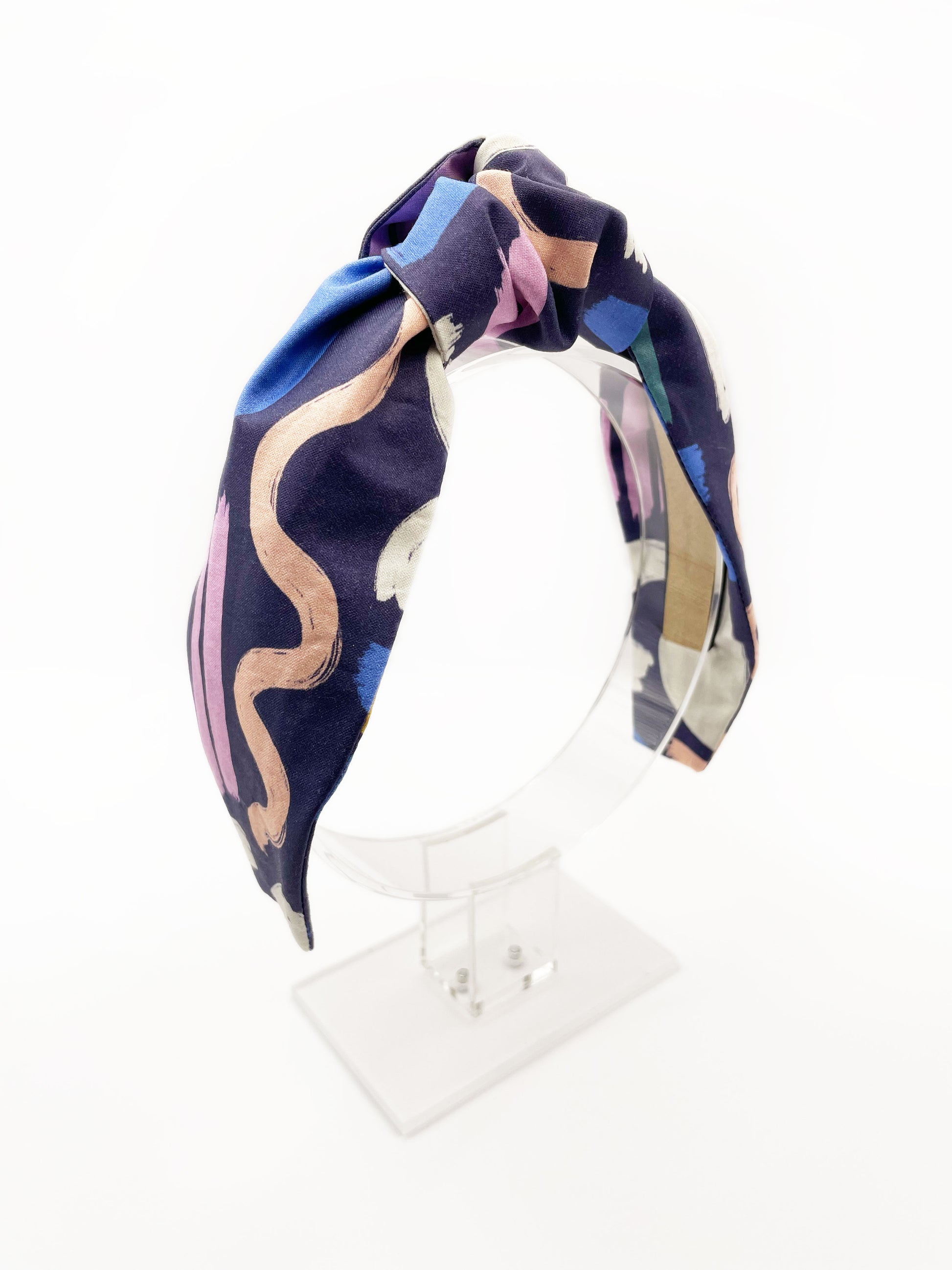 A navy blue knotted headband with a colorful abstract print featuring various doodles. The headband is displayed on an acrylic stand.