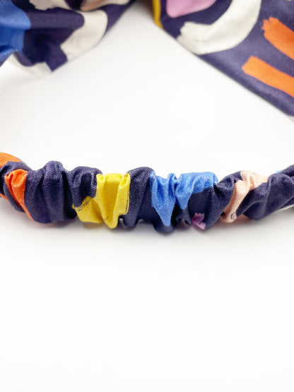 A navy blue twist headband with a colorful abstract print featuring various doodles. The elastic band in the back provides support without slipping.