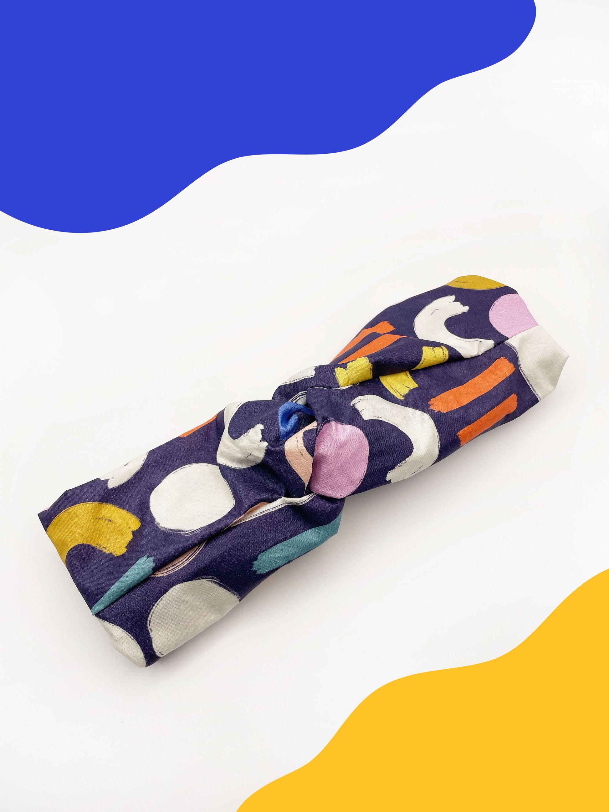 A navy blue twist headband with a colorful abstract print featuring various doodles. The elastic band in the back provides support without slipping.